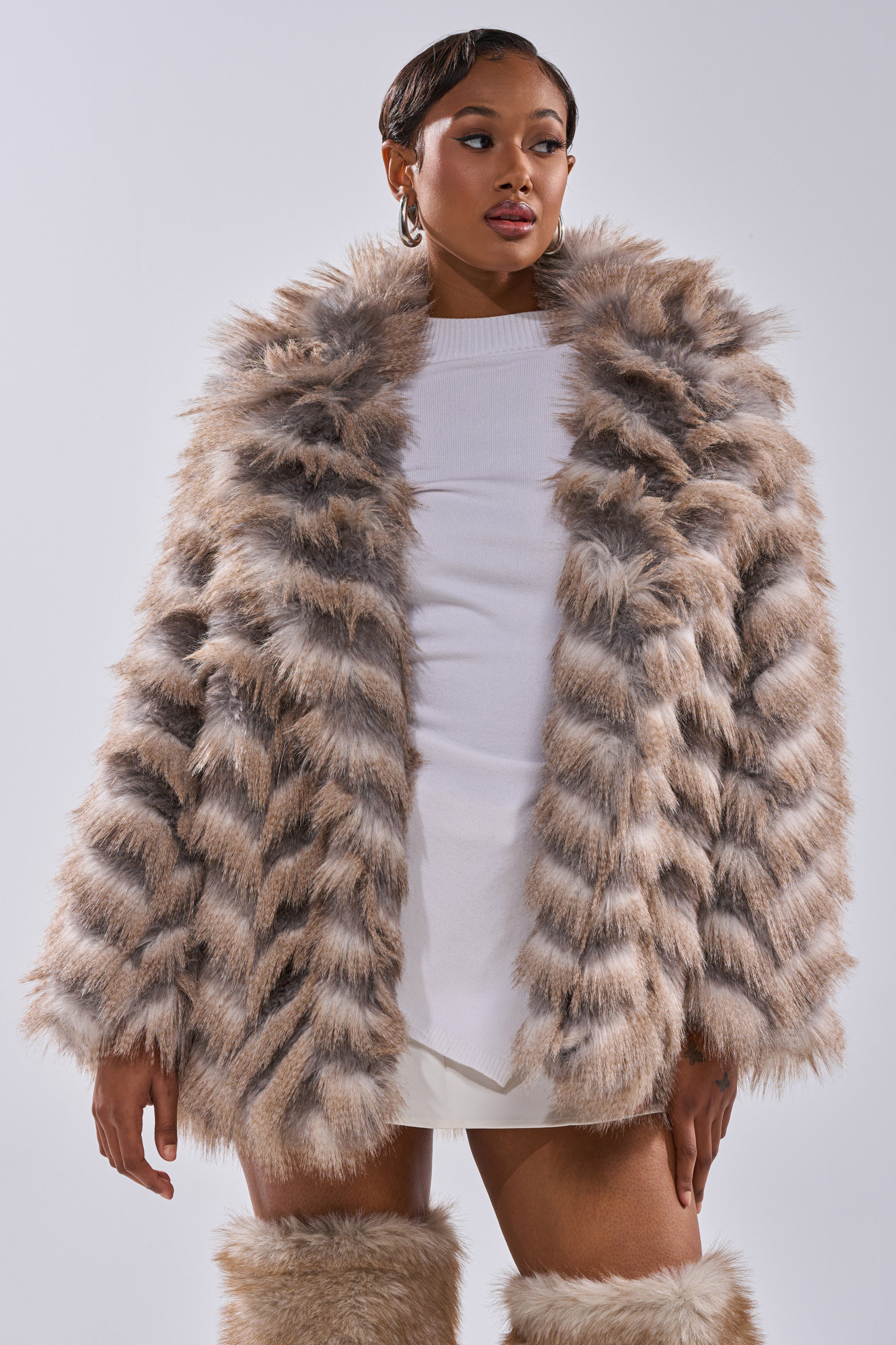 A woman wears the SHOWBIZ BABY FAUX FUR coat in a chevron beige and brown print over a white dress, paired with matching faux fur leg warmers and gold hoop earrings, set against a light background.
