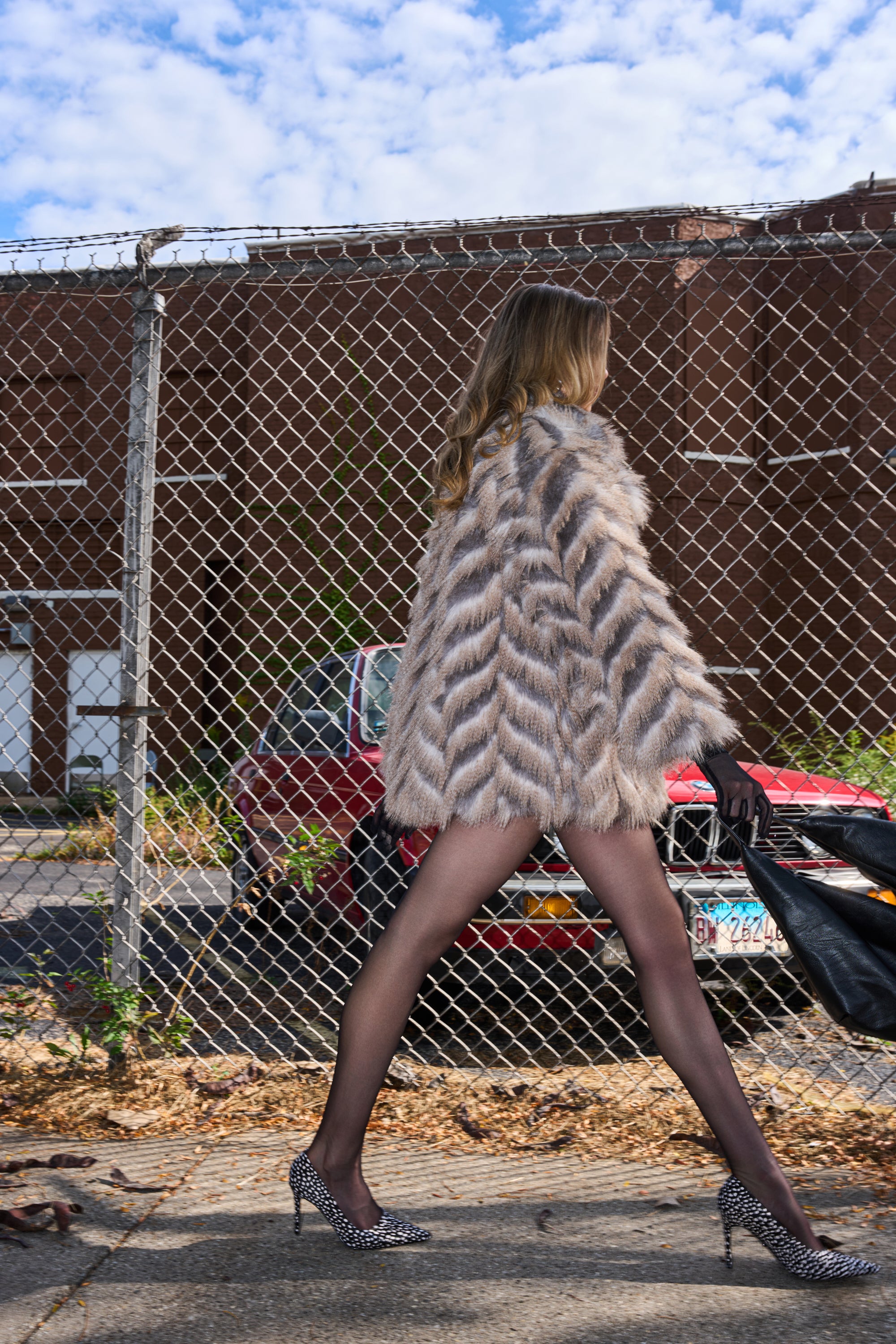 Wearing SHOWBIZ BABY FAUX FUR, sheer black tights, and animal print heels, a woman walks by a chain-link fence with a red car and brick building in the background under partly cloudy skies.