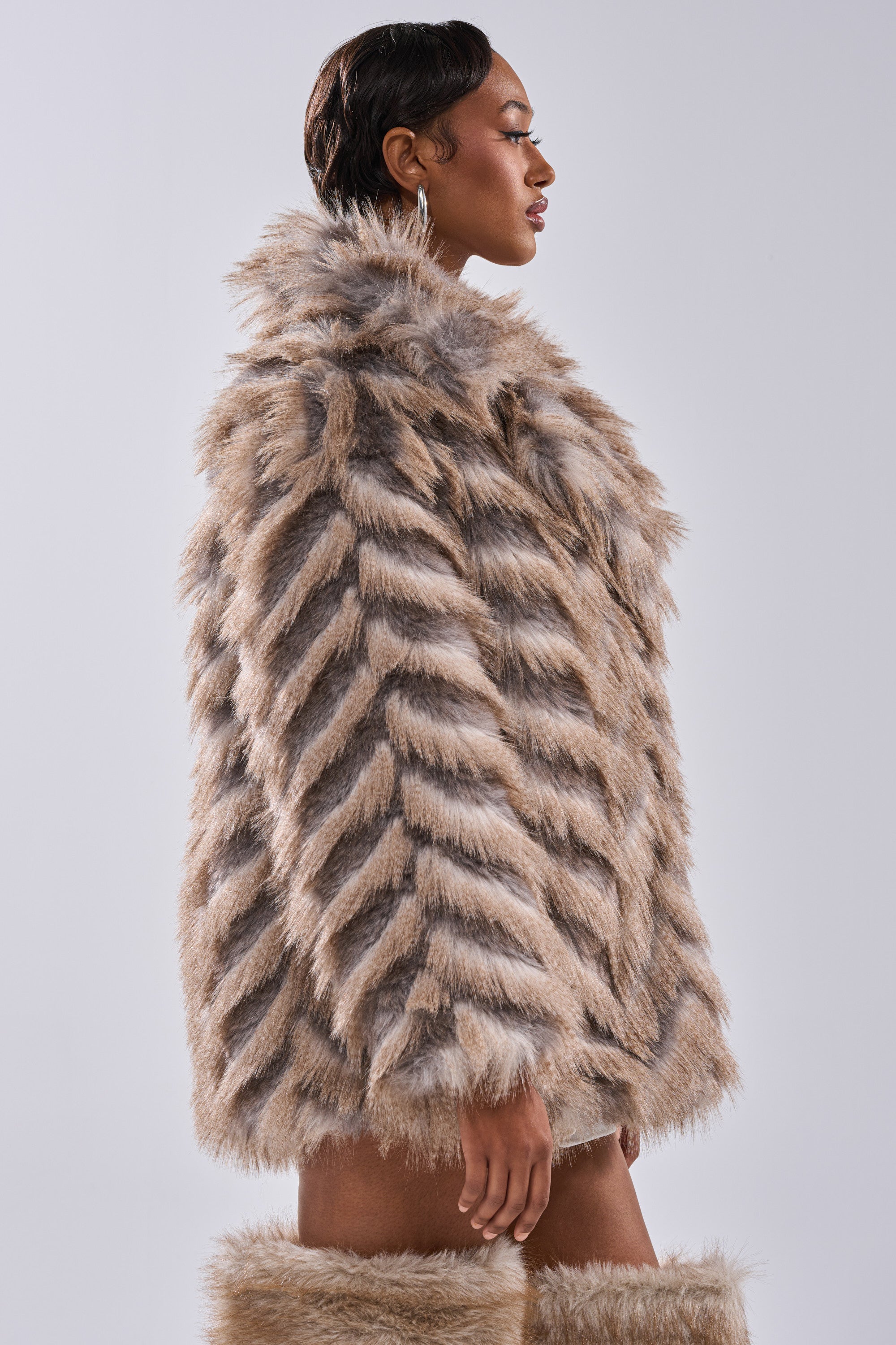 A woman in the SHOWBIZ BABY FAUX FUR chevron print coat and matching boots poses in profile against a light gray background, wearing large hoop earrings and her hair styled neatly.