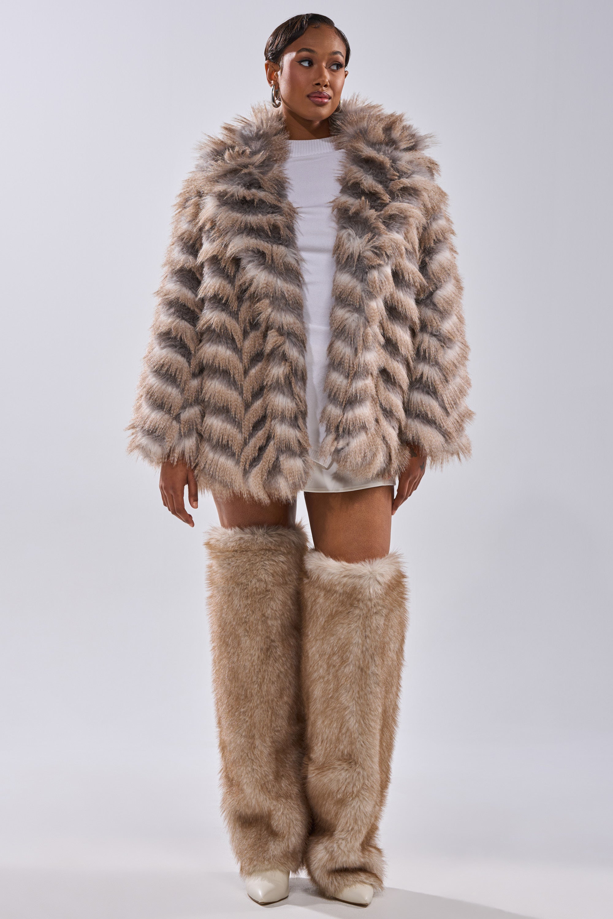 A person poses confidently in the SHOWBIZ BABY FAUX FUR— a striped chevron-print coat with matching furry leg warmers, paired with a short white dress and white shoes against a plain background. They wear their hair short.