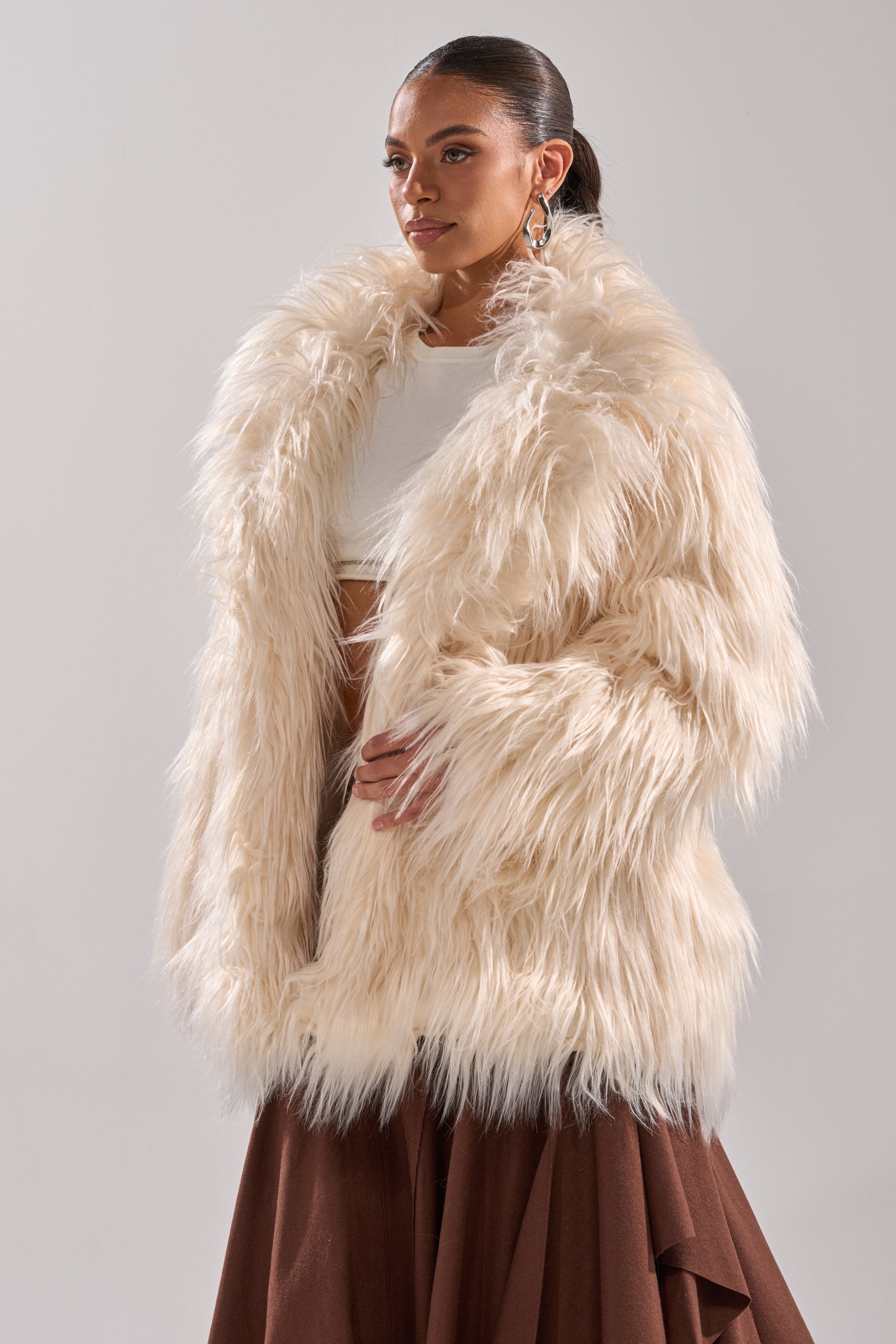A woman stands against a light gray background wearing the PARK CITY FAUX FUR COAT over a white crop top and long brown skirt. Her hair is pulled back, and she completes the look with large hoop earrings.