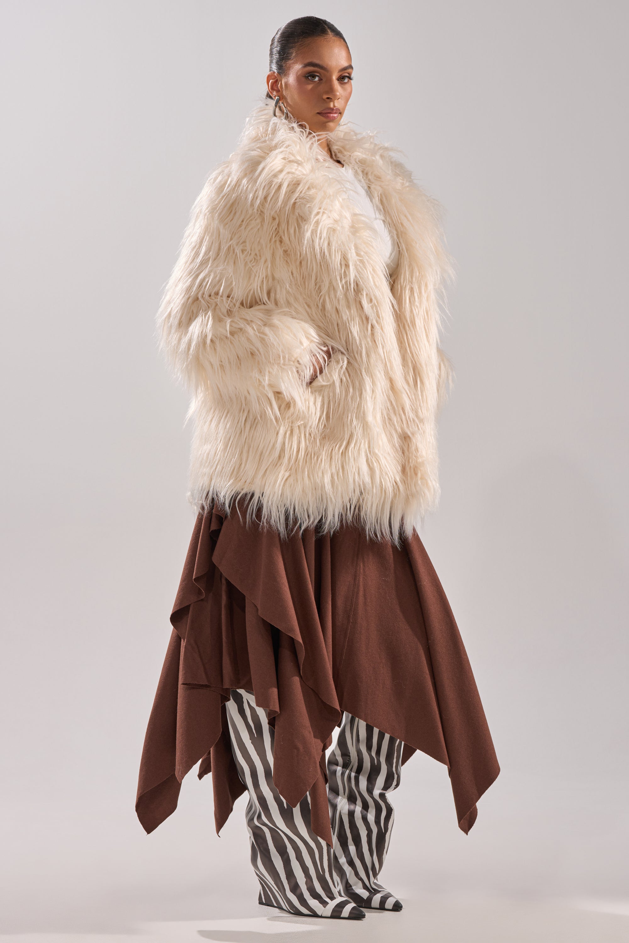 A woman poses in the PARK CITY FAUX FUR COAT, featuring an oversized fit and satin lining, styled with a brown asymmetrical skirt and black-and-white striped pants against a plain light background.