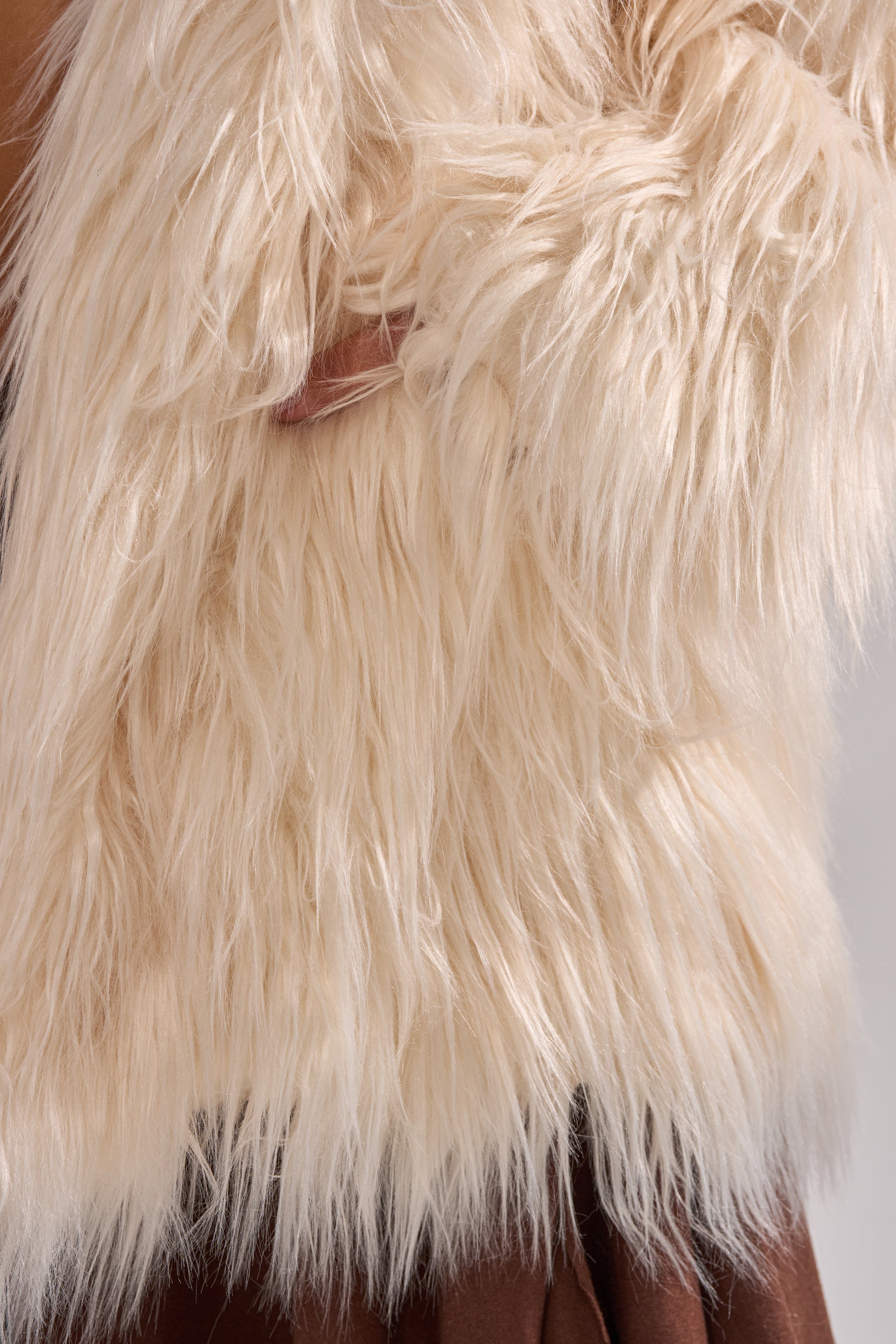 A person wears the PARK CITY FAUX FUR COAT, an oversized, shaggy cream layer over a brown outfit, their hand nestled in its plush texture.