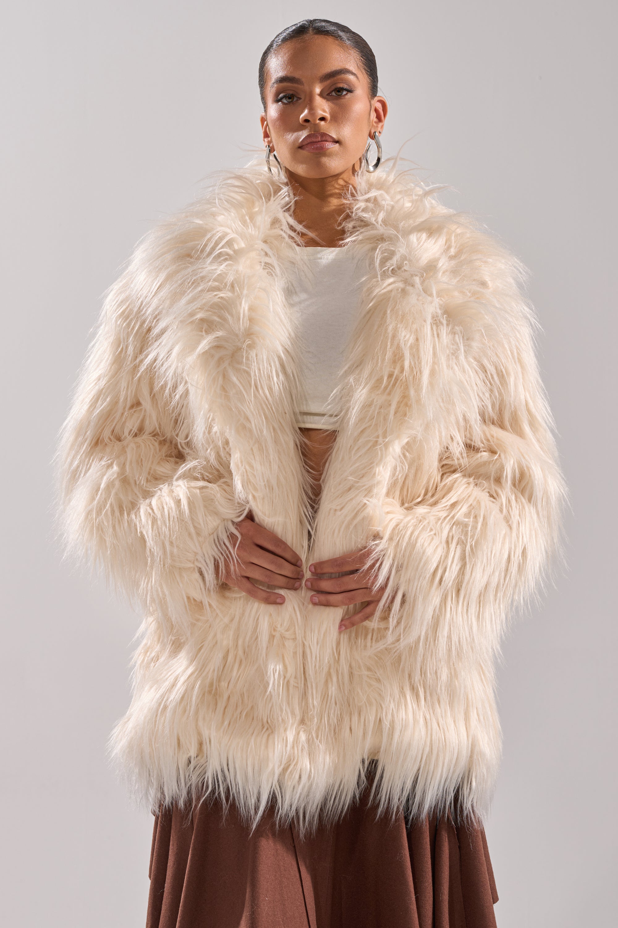 A person models the PARK CITY FAUX FUR COAT over a white crop top and brown skirt, standing against a plain gray background.