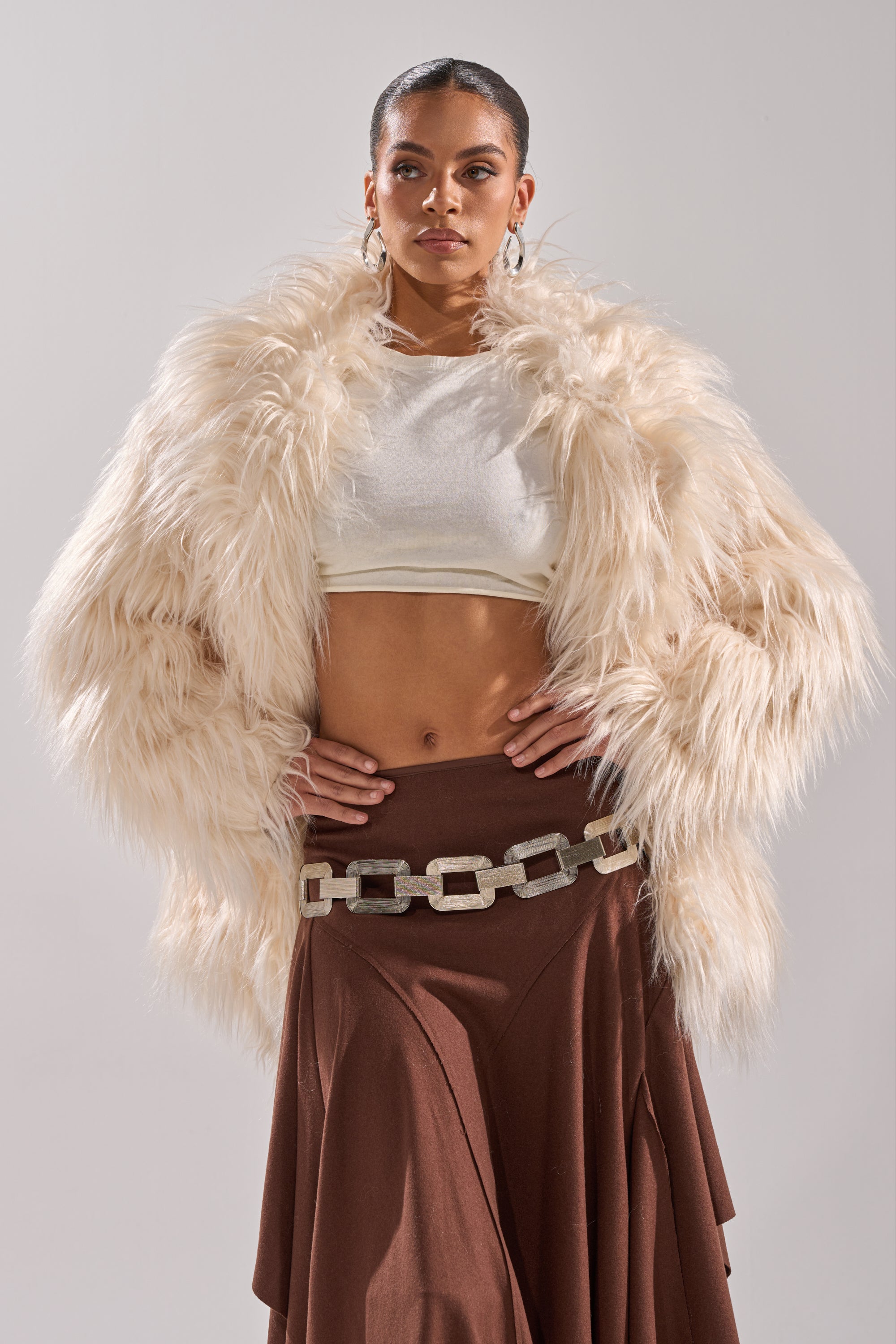 A woman poses confidently in the PARK CITY FAUX FUR COAT with satin lining, styled over a white crop top, brown asymmetrical skirt, and chunky chain belt against a plain light background.