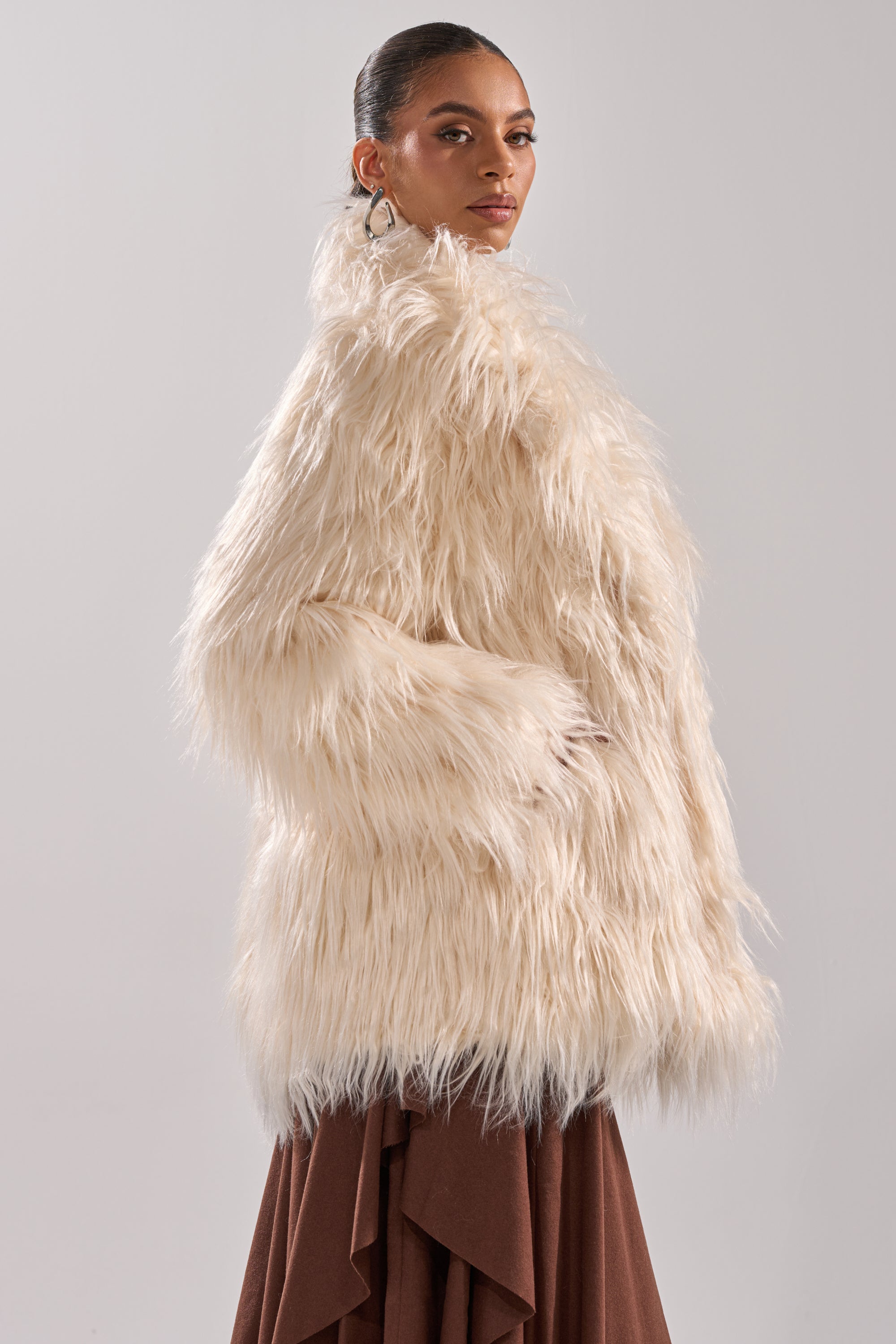 A woman in the PARK CITY FAUX FUR COAT, cream-colored and long with a satin lining, stands against a plain light background over a brown skirt, looking over her shoulder confidently.