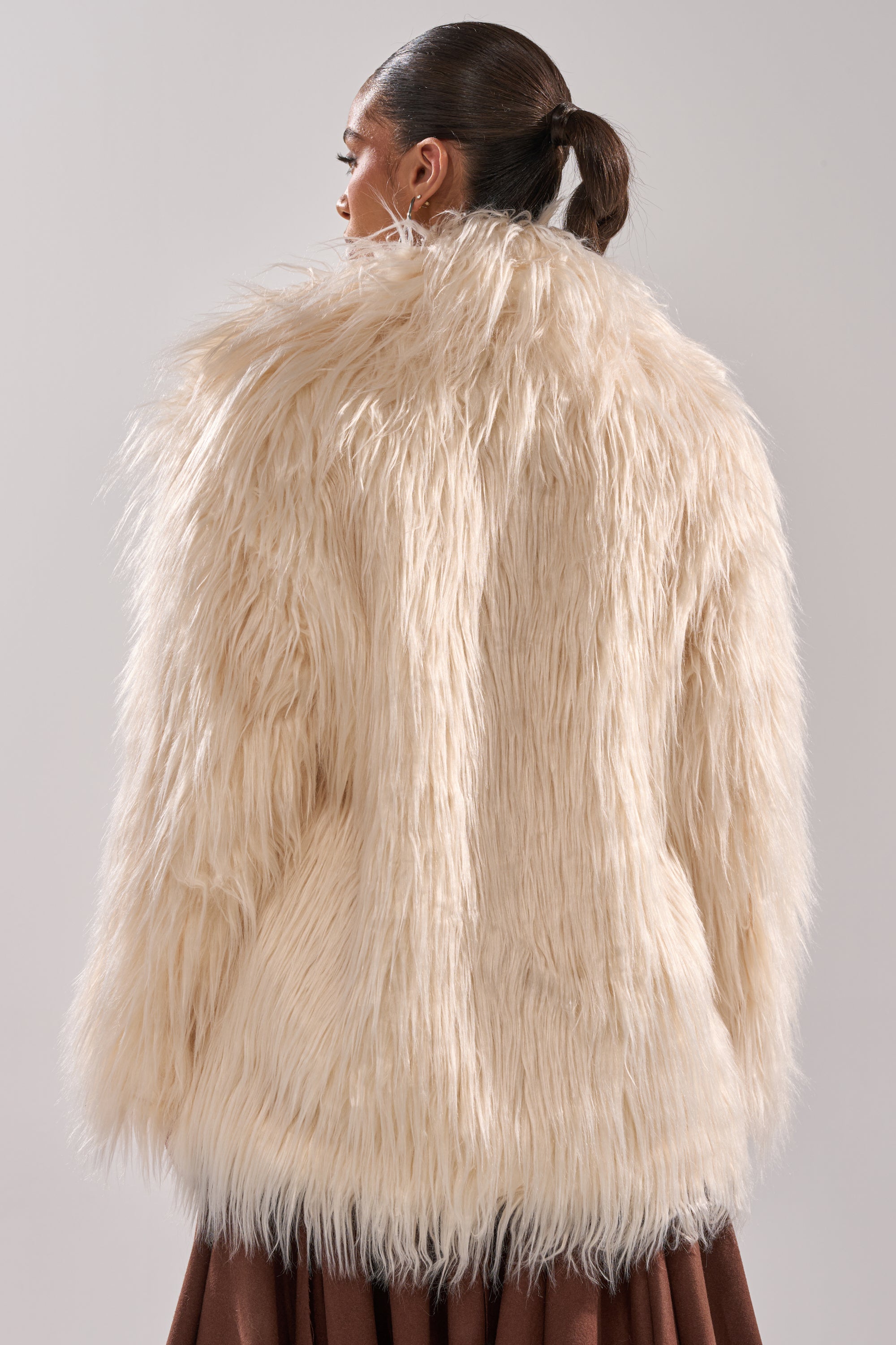 A woman with dark hair in a ponytail is seen from the back, wearing the PARK CITY FAUX FUR COAT in off-white with a cozy satin lining and a brown skirt, standing against a plain light gray background.