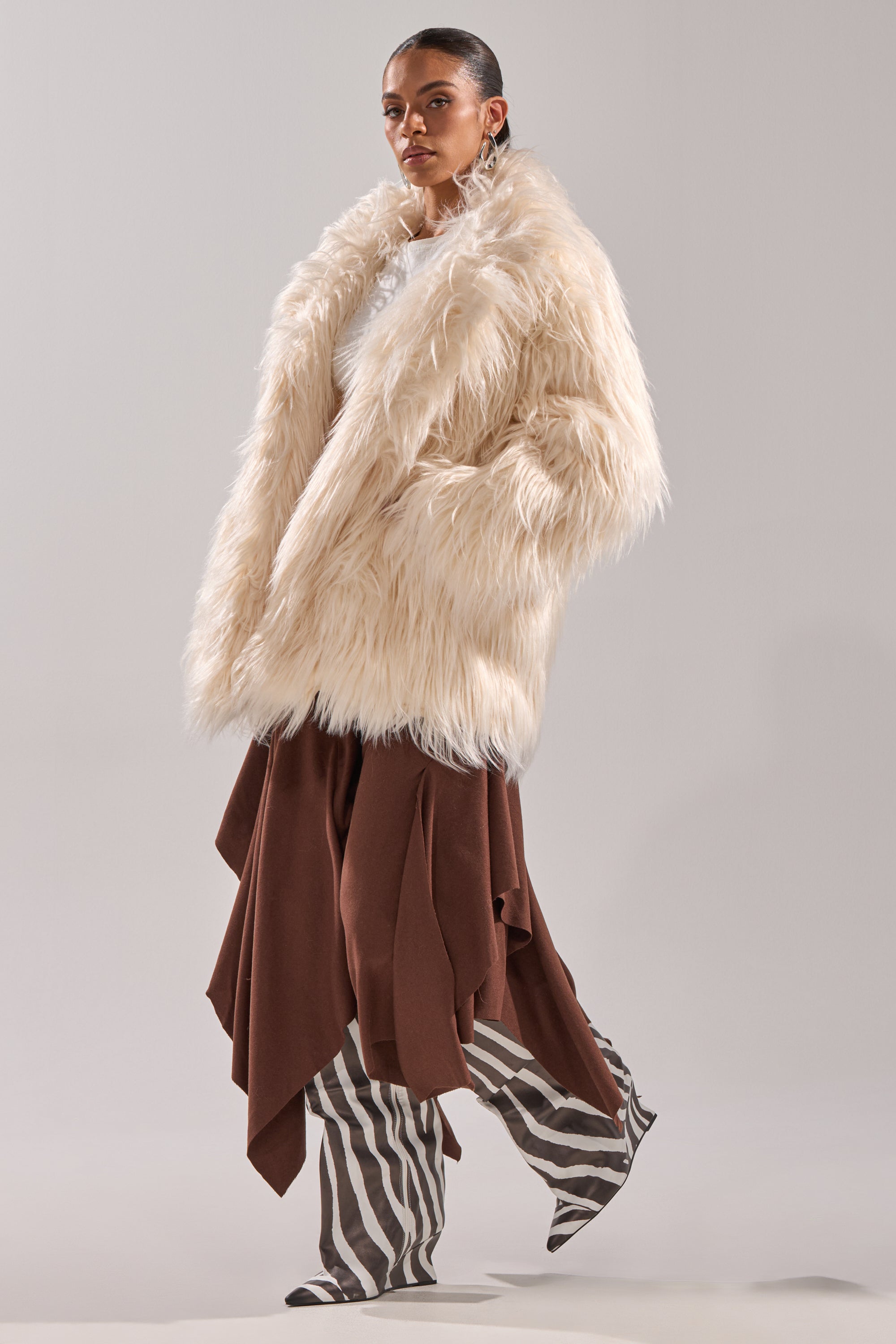 A woman poses confidently in the PARK CITY FAUX FUR COAT, a cream faux fur layer, styled over a brown skirt and striped pants against a plain light gray background.
