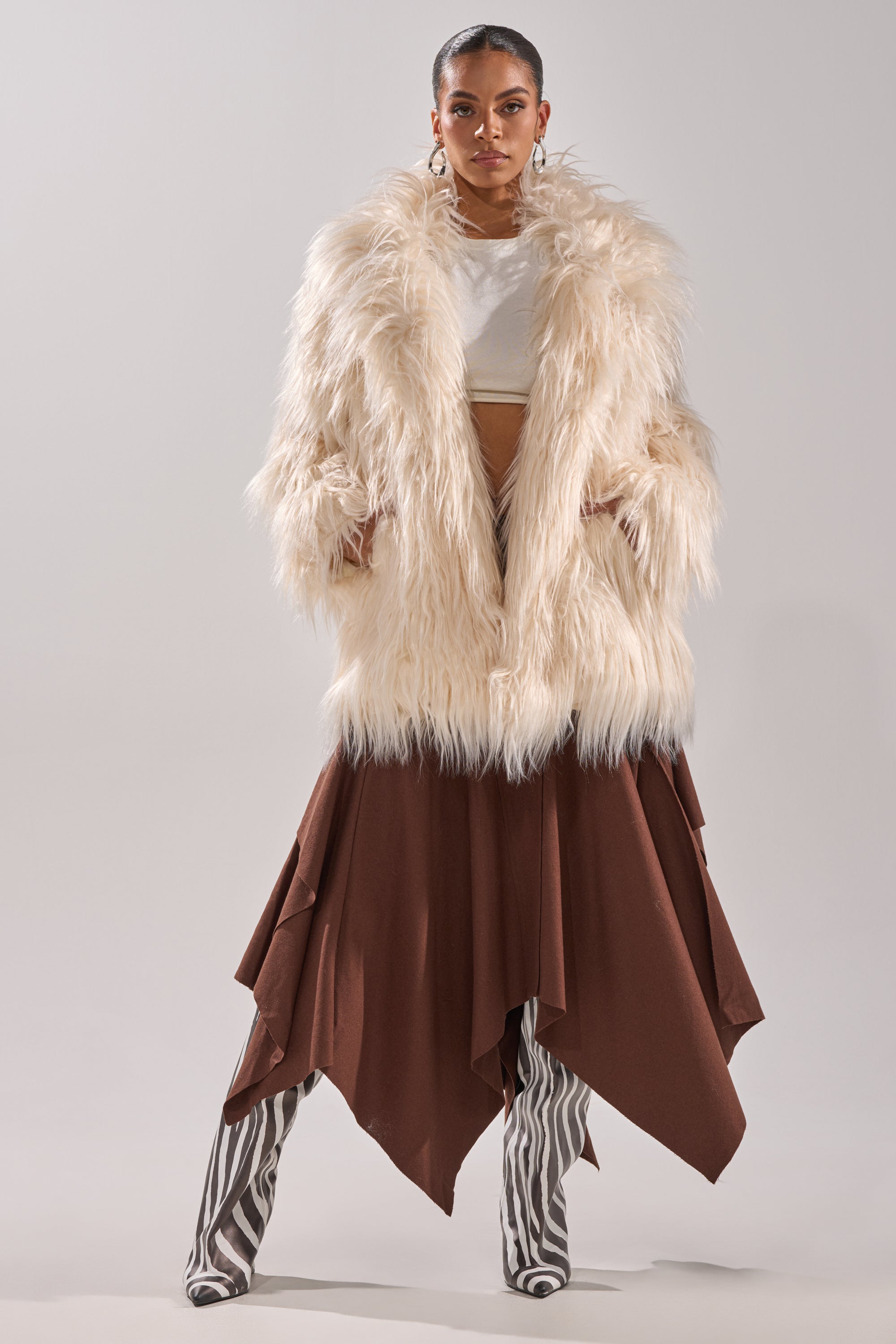 A woman stands confidently in the PARK CITY FAUX FUR COAT over a white top, layered brown skirt, and bold black-and-white zebra-striped boots against a plain light background.