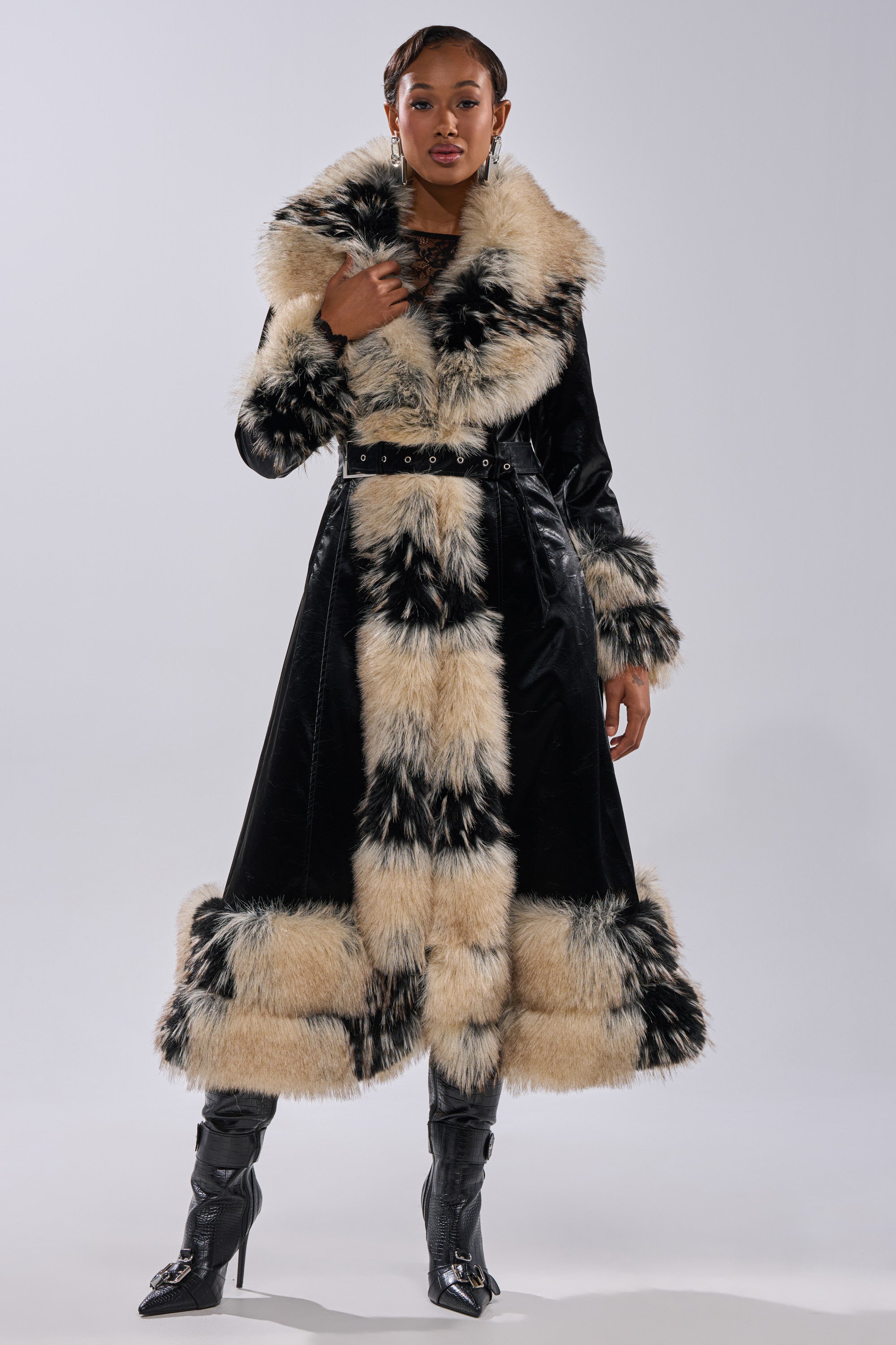 A woman wears the VIVIAN FUR TRIM TRENCH IN LIQUID VELVET, a long black trench featuring thick beige and black faux fur on the collar, cuffs, hem, and belt, styled with pointed black boots against a plain light background.