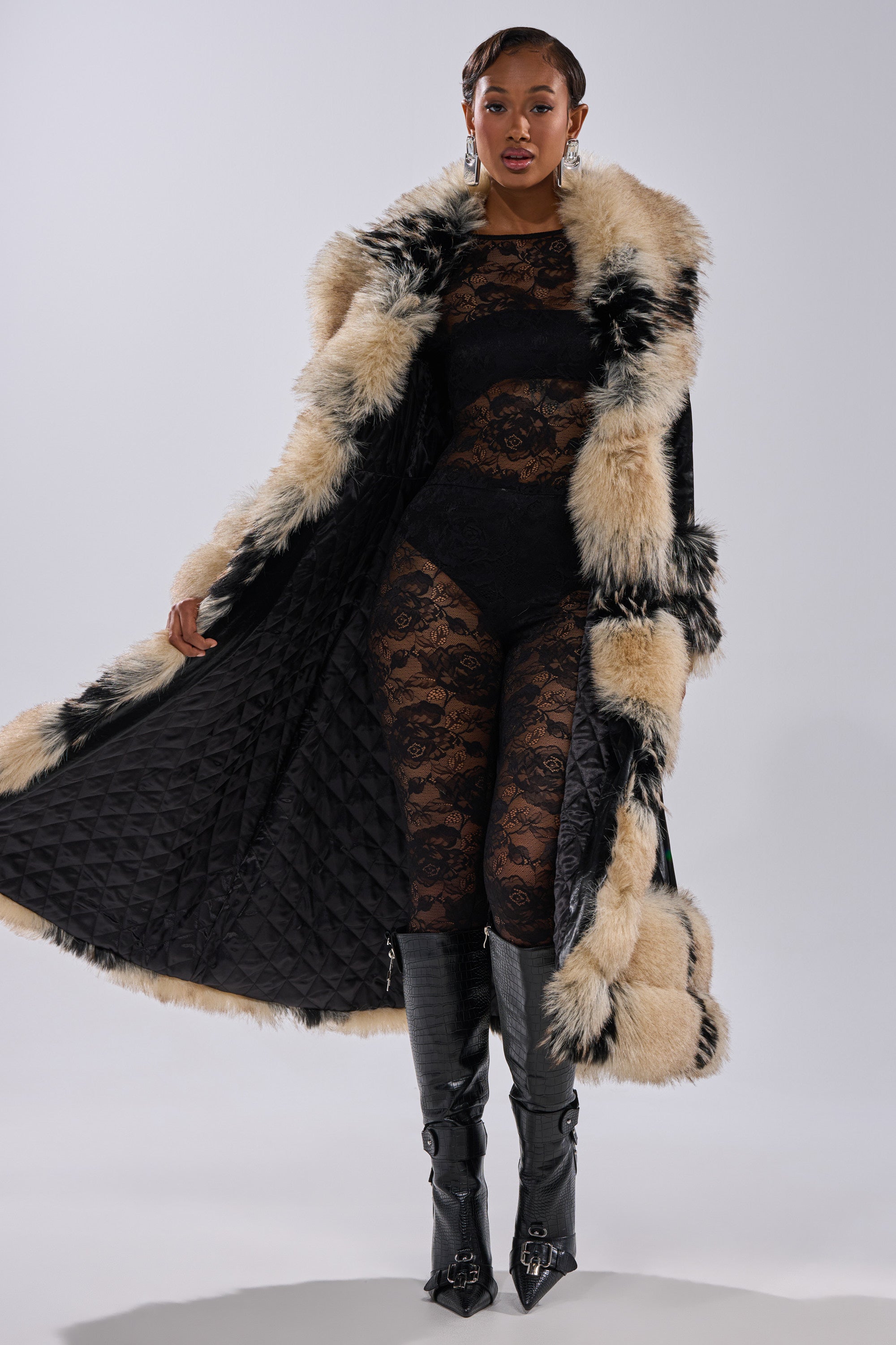 A woman models the VIVIAN FUR TRIM TRENCH IN LIQUID VELVET over a sheer black lace bodysuit and knee-high black boots, exuding confidence against a plain light background.