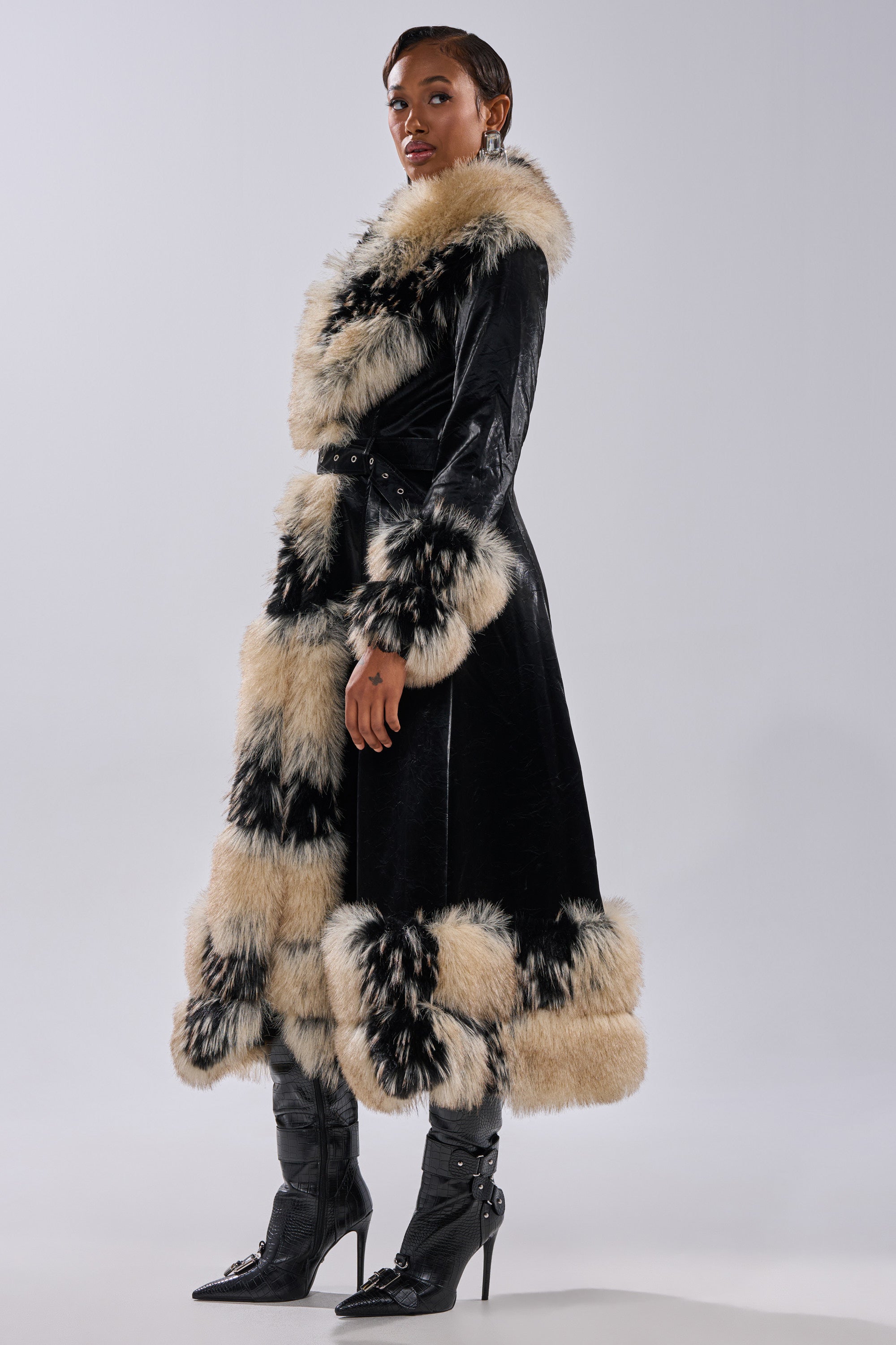 A person wears the VIVIAN FUR TRIM TRENCH IN LIQUID VELVET—a long, black velvet jacket with wide cream-and-black fur trim—paired with black heeled boots, standing against a plain light background.