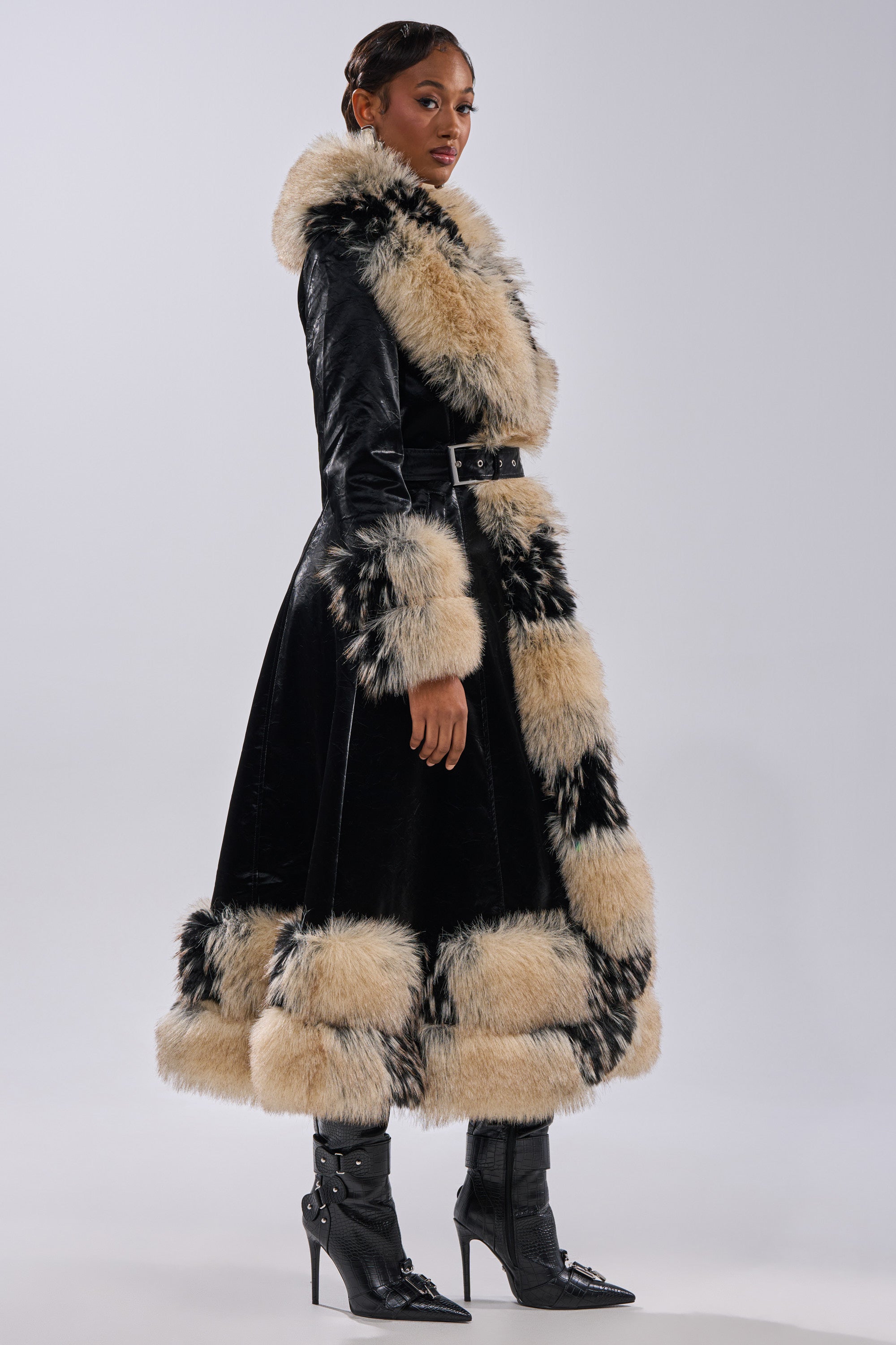 A person models the VIVIAN FUR TRIM TRENCH IN LIQUID VELVET, a long black coat with thick beige and black fur trim on the collar, cuffs, hem, and front. They pair it with pointed black heeled boots against a plain light background.
