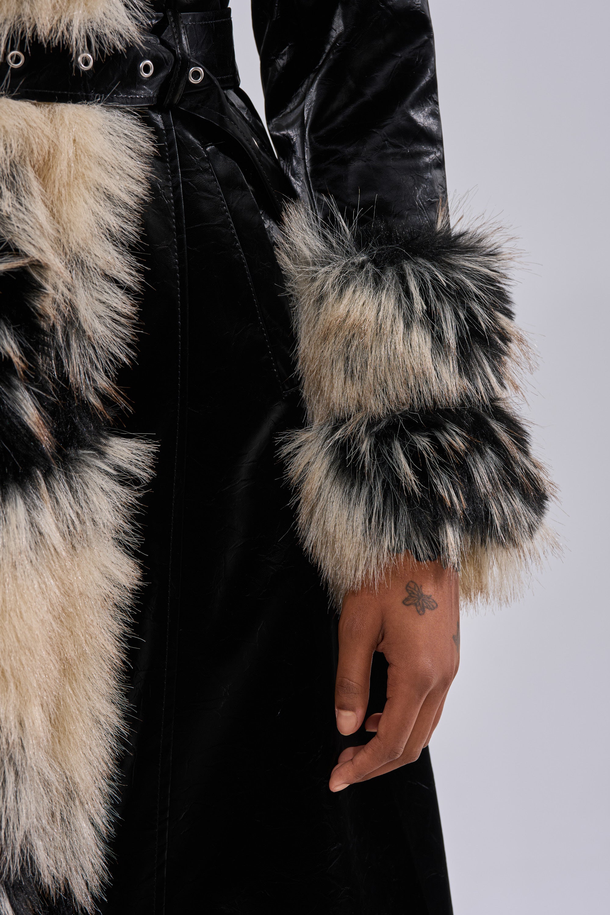 A close-up of a person wearing the VIVIAN FUR TRIM TRENCH IN LIQUID VELVET, with black and white fur trim. The person's hand, displaying a small butterfly tattoo, is visible beside the coat's sleeve.