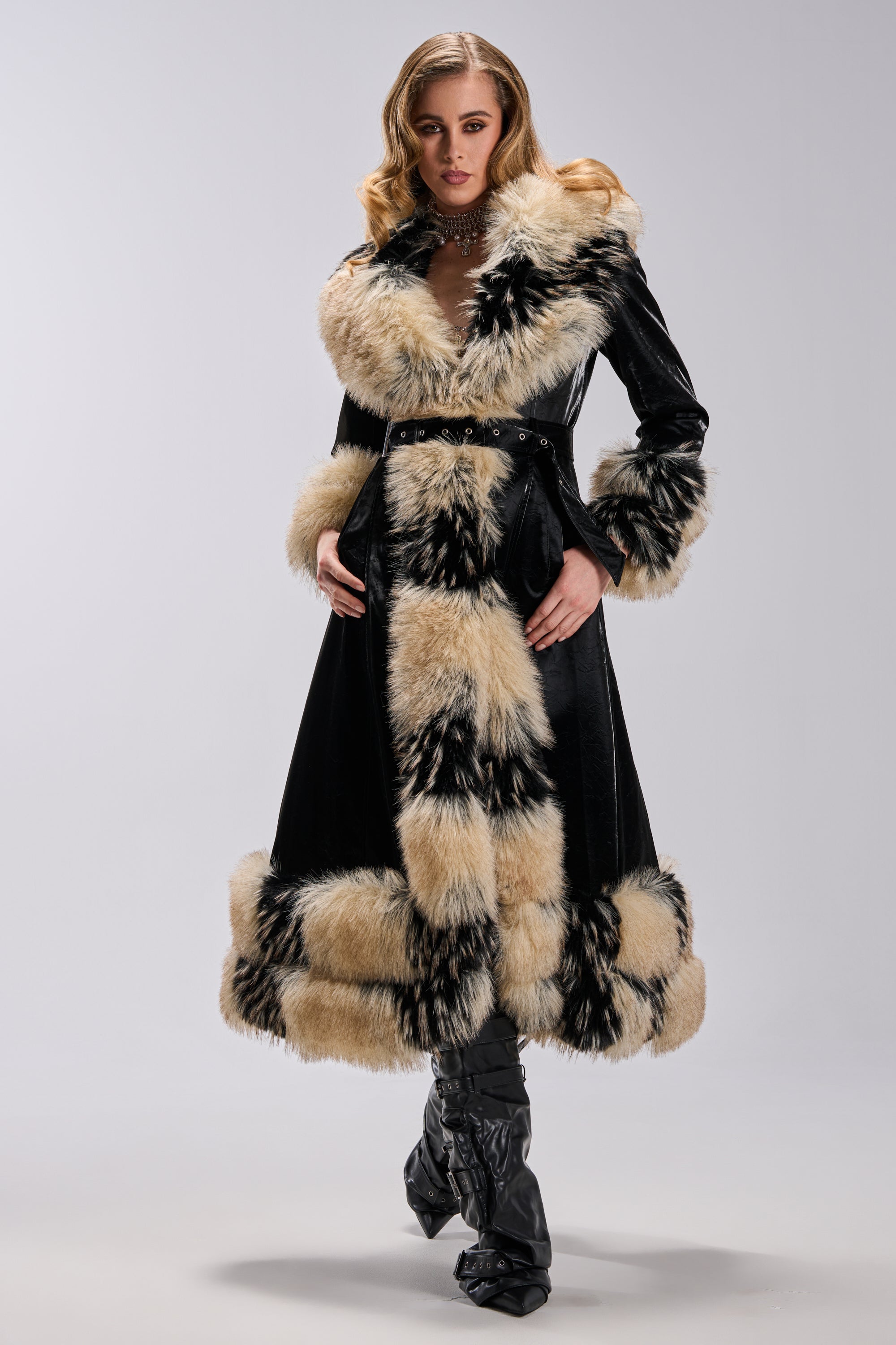A woman models the VIVIAN FUR TRIM TRENCH IN LIQUID VELVET, a long black coat with thick beige and black faux fur on the collar, cuffs, hem, and front, styled with black heeled boots against a plain light background.
