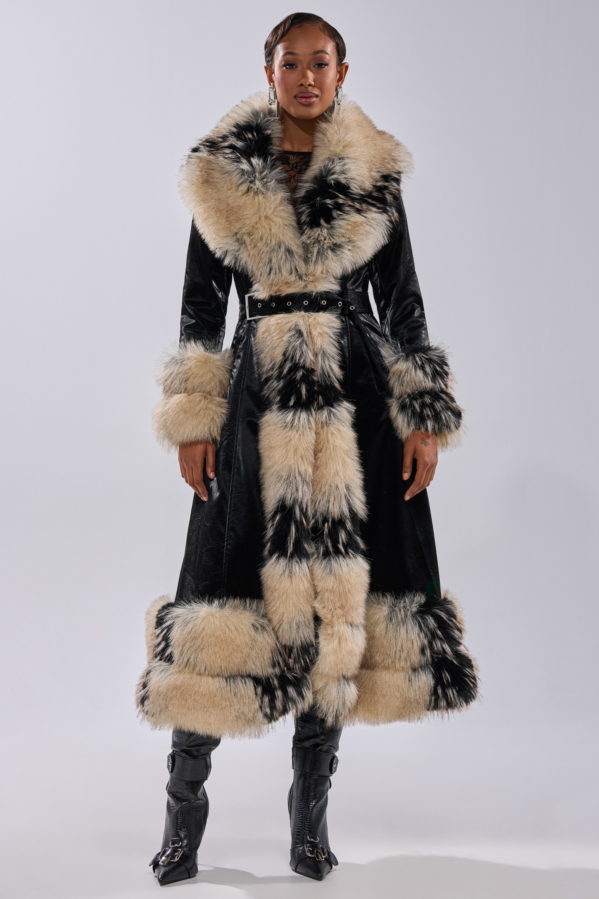 A woman faces forward in the VIVIAN FUR TRIM TRENCH IN LIQUID VELVET, a long black coat with thick cream and black faux fur on the collar, cuffs, hem, and front. She pairs it with black boots and exudes confidence.