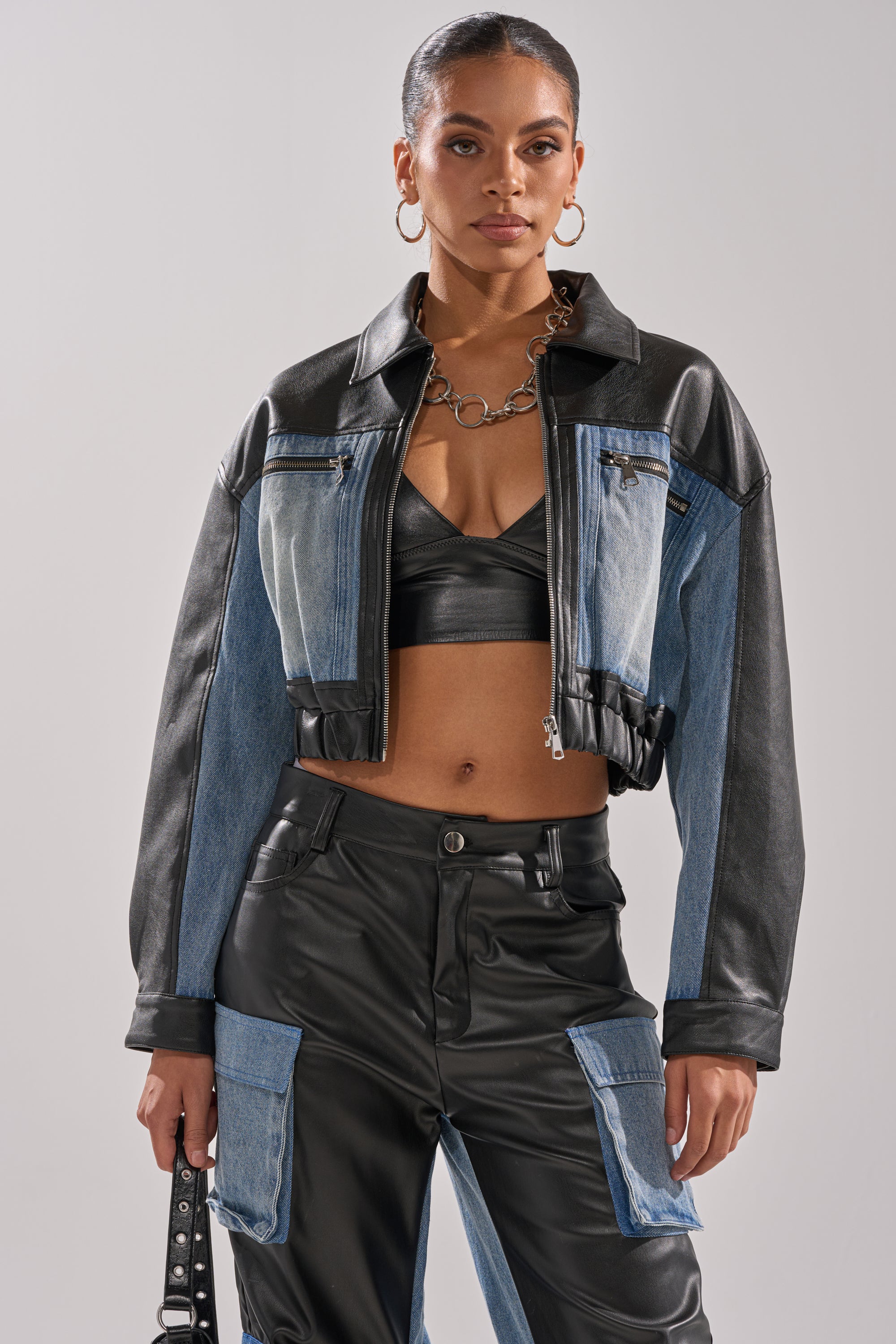 A woman wears the DOWN TIME MIXED MEDIA BOMBER with denim and black faux leather, paired with a black bralette and matching pants. She accessorizes with hoop earrings, a chunky chain necklace, and carries a small handbag.