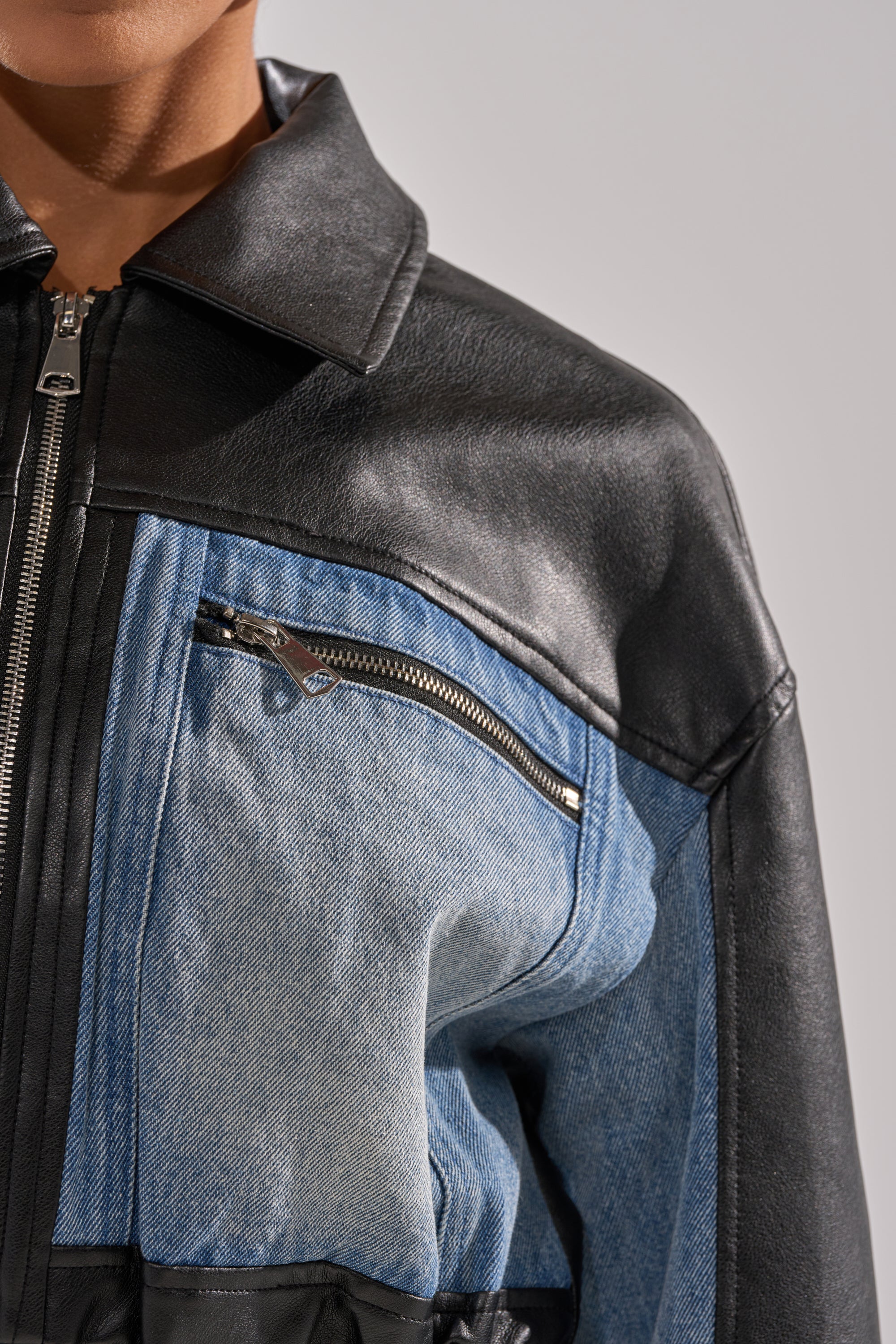 Close-up of a person wearing the DOWN TIME MIXED MEDIA BOMBER, featuring black faux leather and blue denim, with a front zipper and diagonal zippered chest pocket. Only the lower half of the face and upper torso are visible.