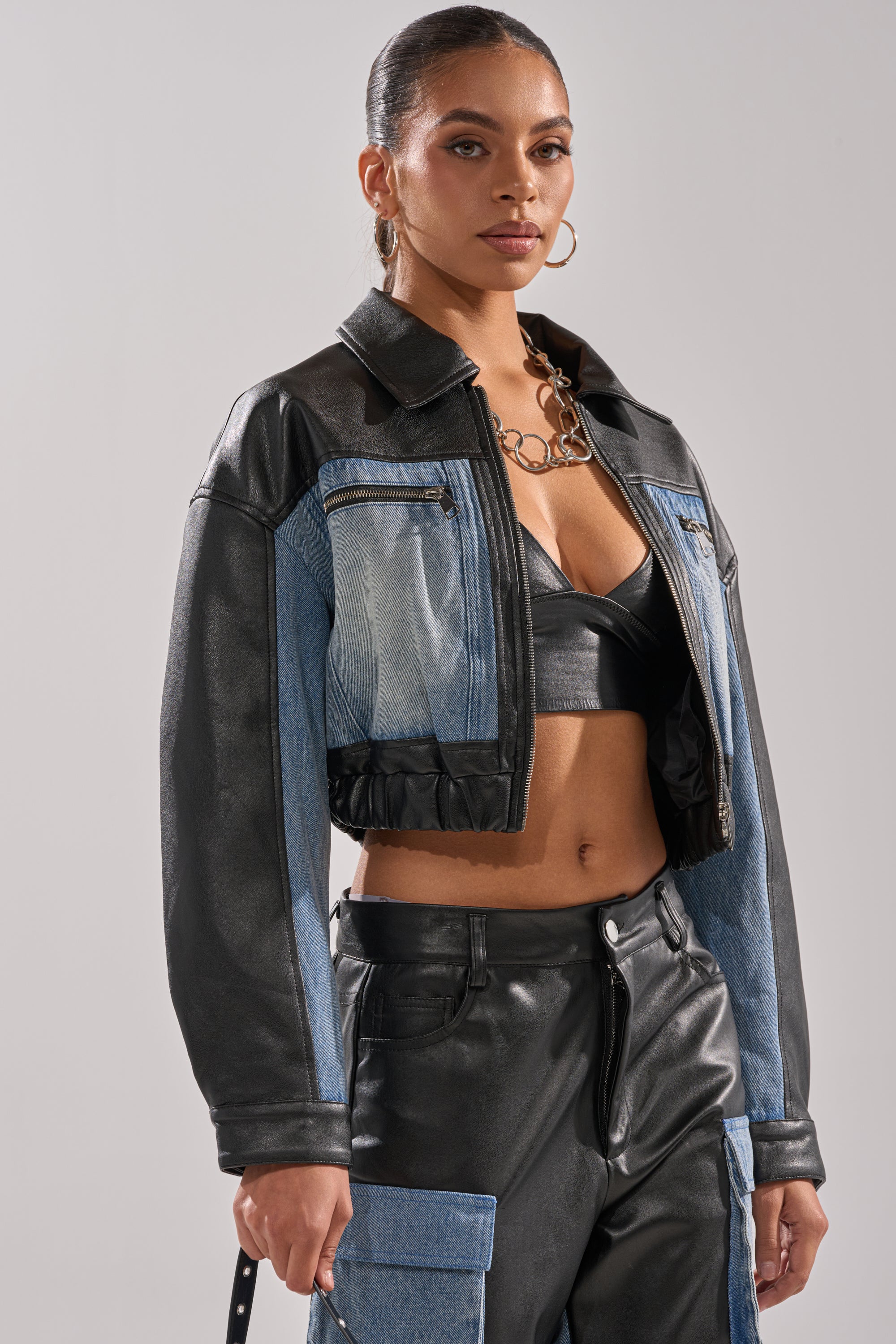 A woman styles the DOWN TIME MIXED MEDIA BOMBER over a black leather bralette and patchwork pants, finishing her streetwear look with gold hoop earrings and a chunky chain necklace.