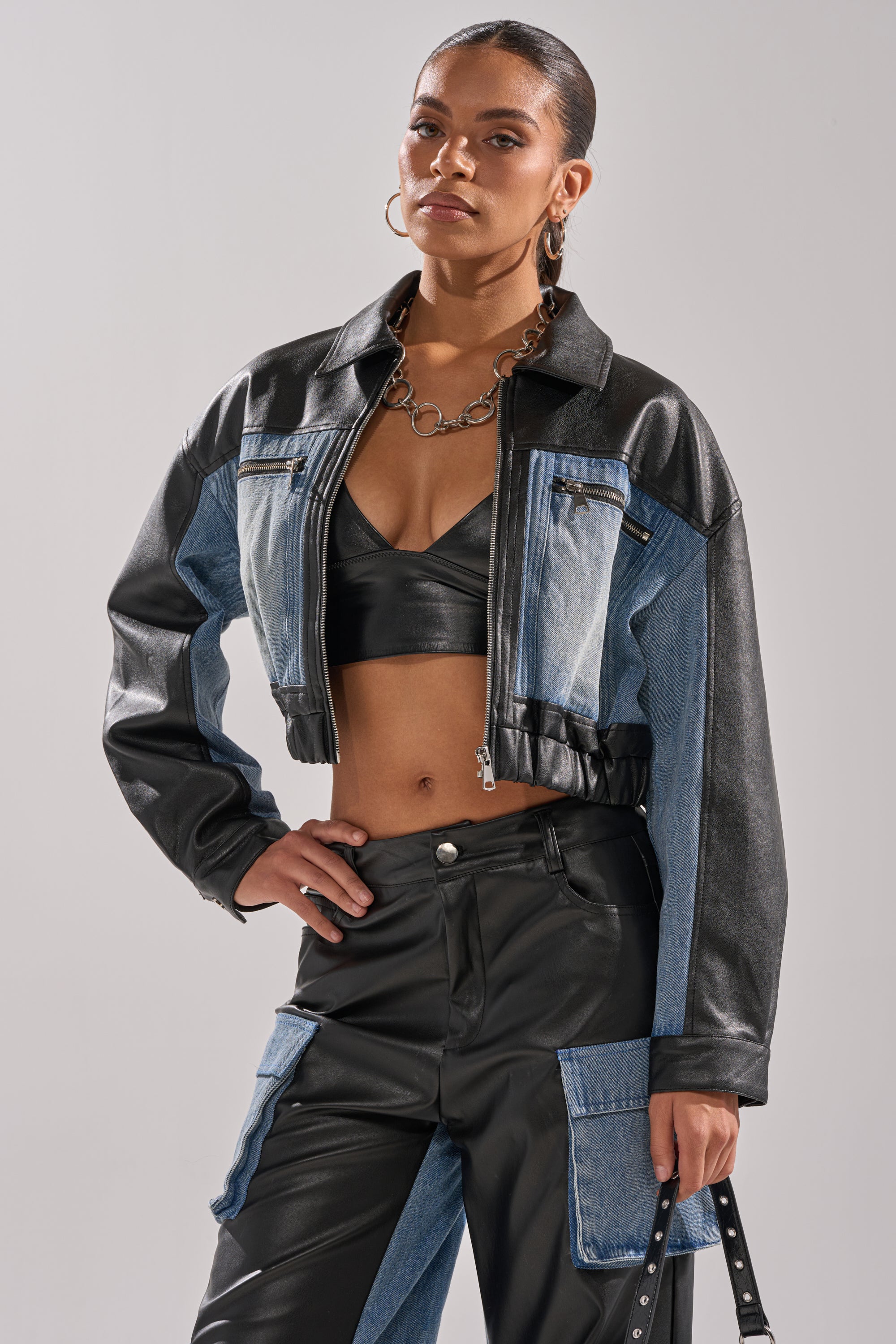 A woman poses confidently in the DOWN TIME MIXED MEDIA BOMBER—a stylish cropped jacket paired with a black bralette and matching patchwork pants. She completes her streetwear look with hoop earrings and a chunky chain necklace.