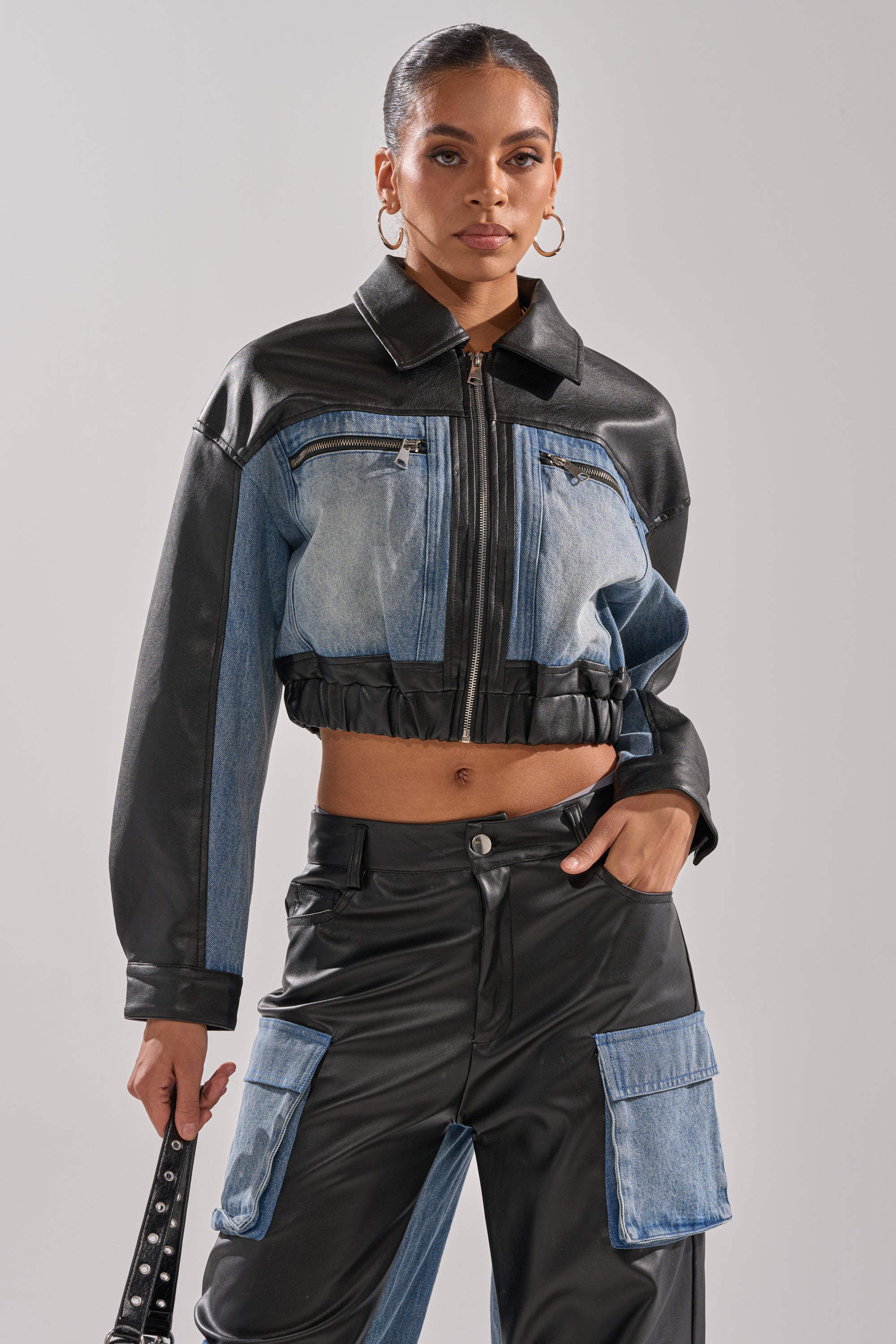 A woman styles the DOWN TIME MIXED MEDIA BOMBER, a faux leather zip-up jacket, with matching high-waisted pants featuring denim pockets, hoop earrings, and a sleek bun for a streetwear look.