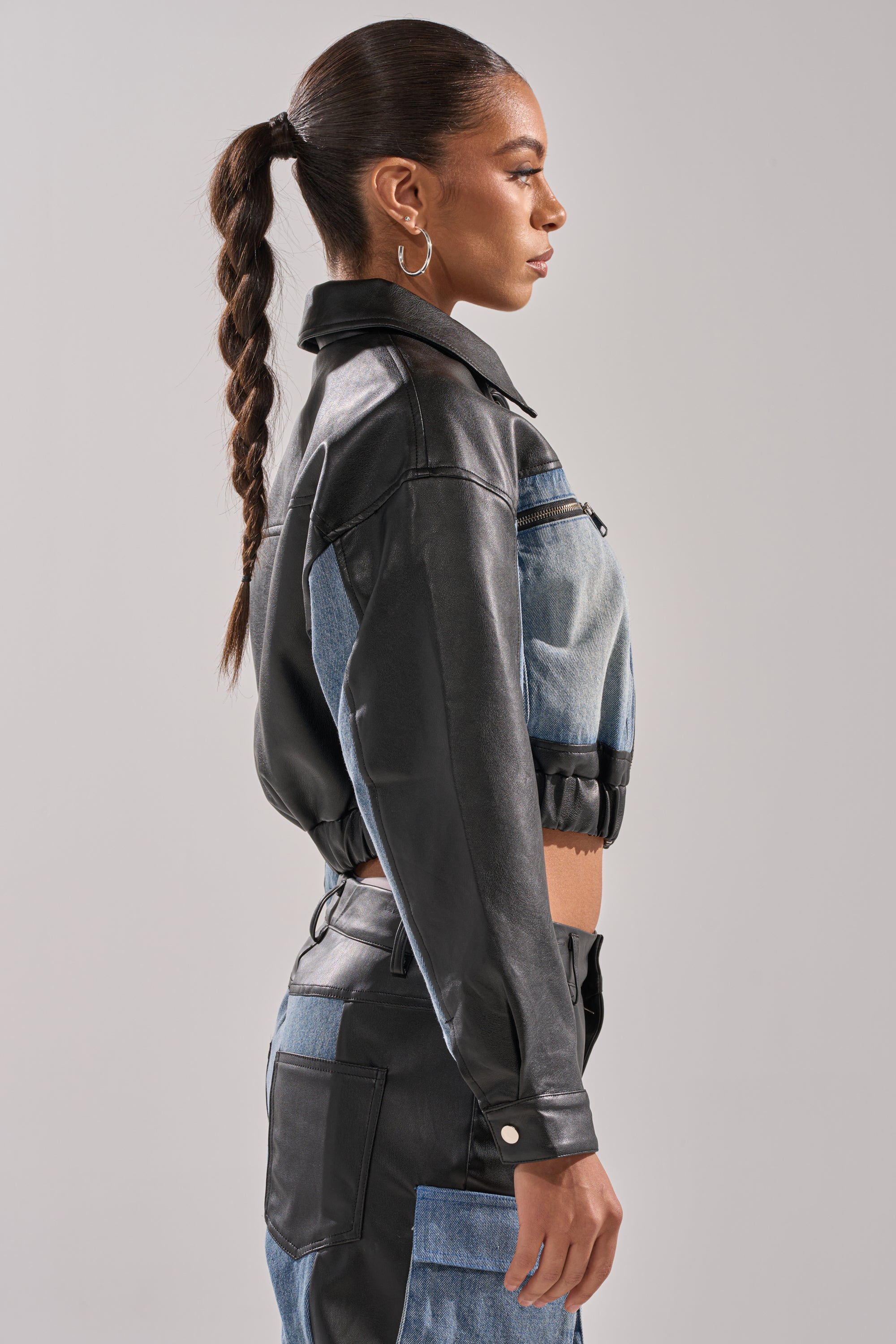 A woman with a braided ponytail models the DOWN TIME MIXED MEDIA BOMBER—a cropped black and denim jacket with matching high-waisted pants—standing in profile against a plain light gray background.