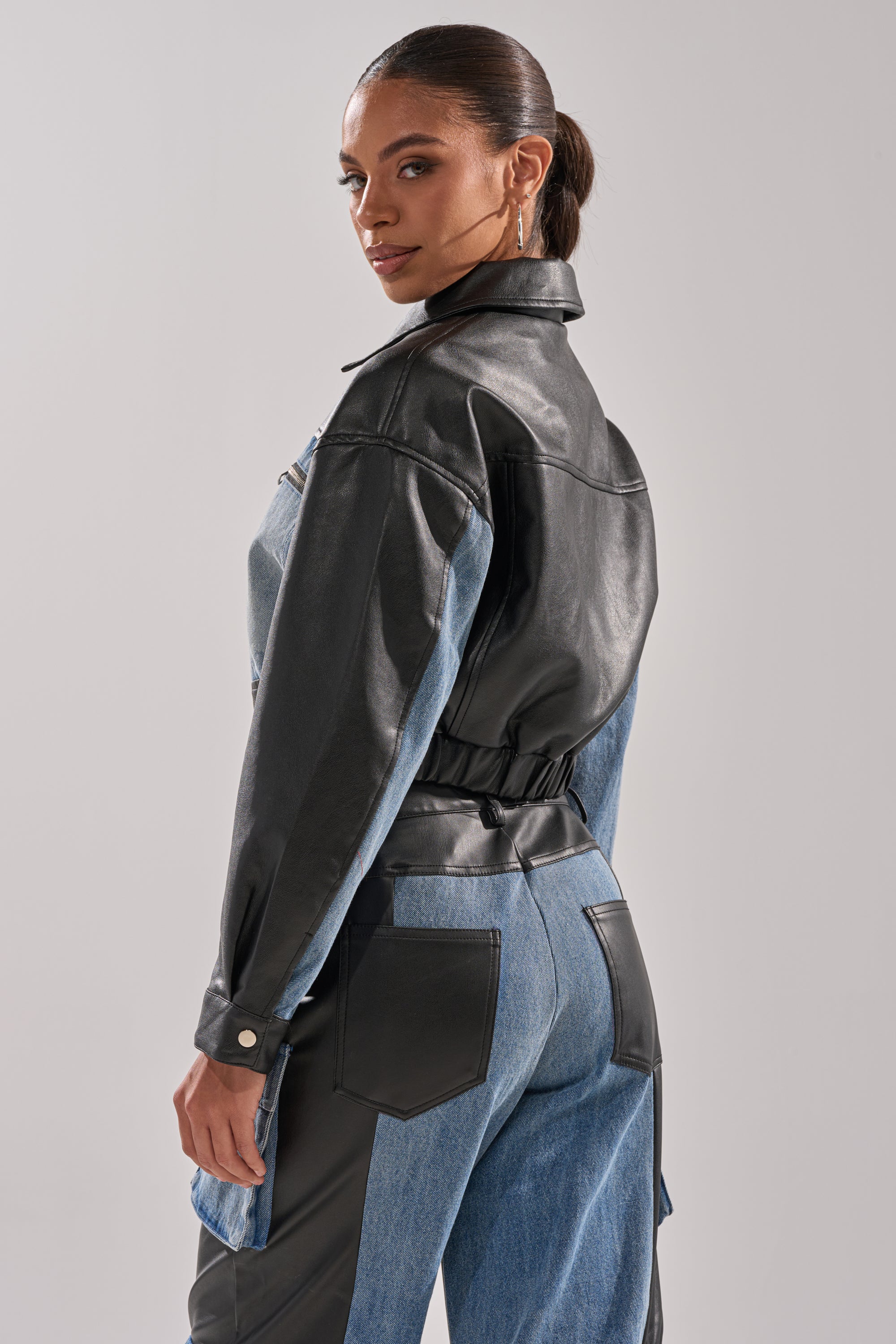 A woman stands turned slightly to the side, looking over her shoulder. She wears the DOWN TIME MIXED MEDIA BOMBER and matching pants with black faux leather accents and patch pockets, creating a sleek streetwear vibe.