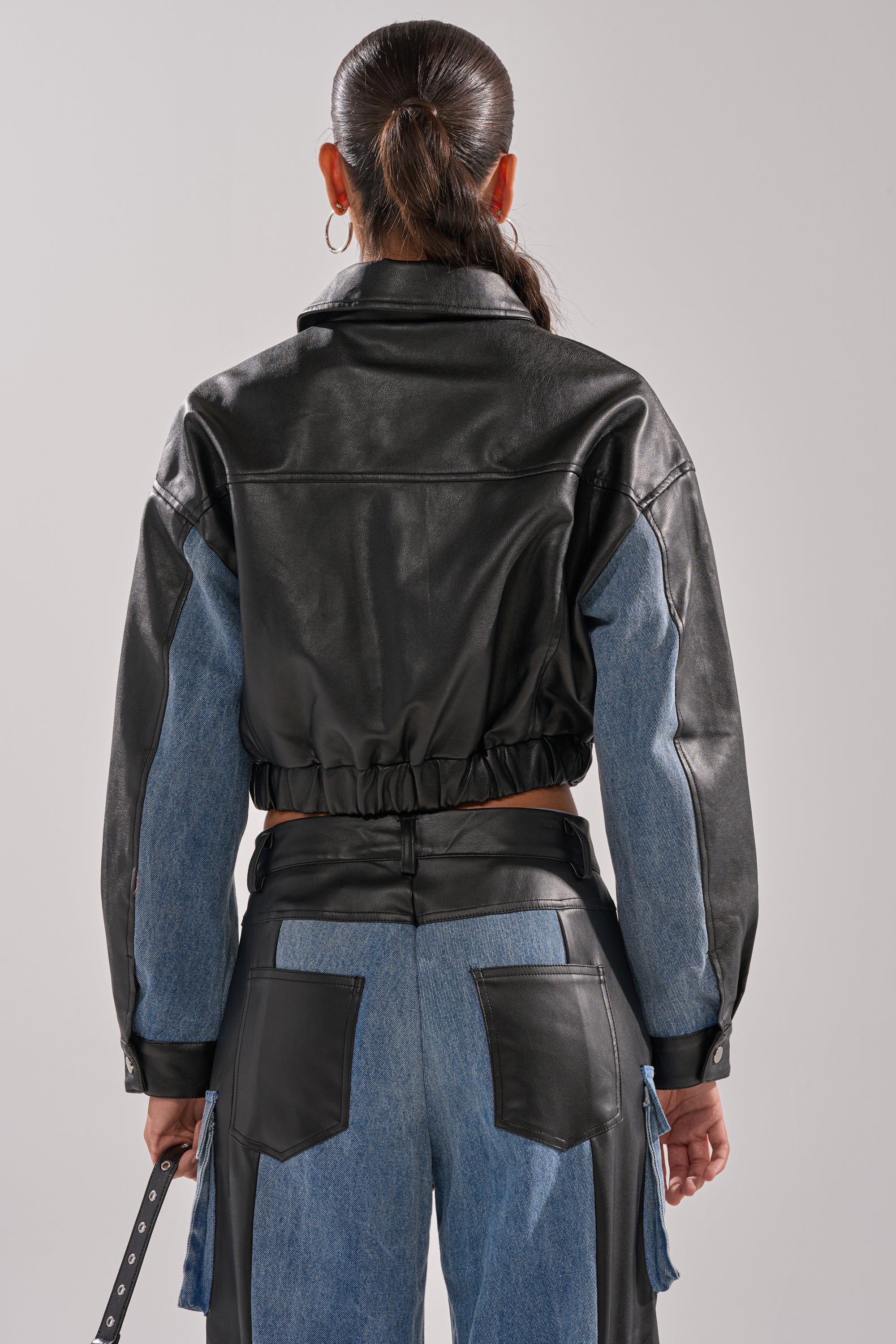 A person with long dark hair in a ponytail wears the DOWN TIME MIXED MEDIA BOMBER and denim pants with black leather patches, shown from the back against a plain light background.