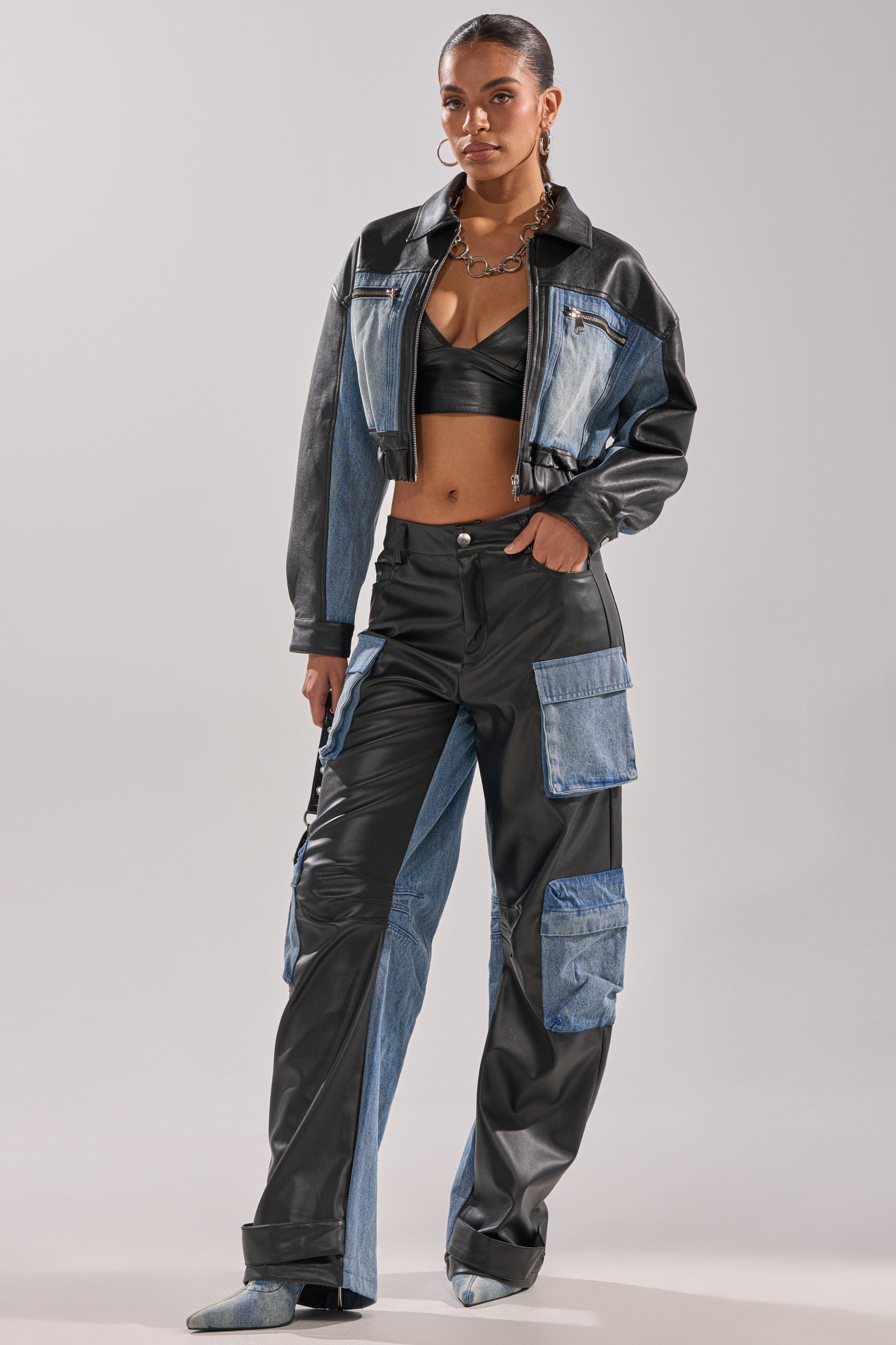 A woman stands confidently in the DOWN TIME MIXED MEDIA BOMBER, styled with a black bralette and high-waisted denim and black leather patchwork pants, pointed-toe shoes, and a chunky necklace.