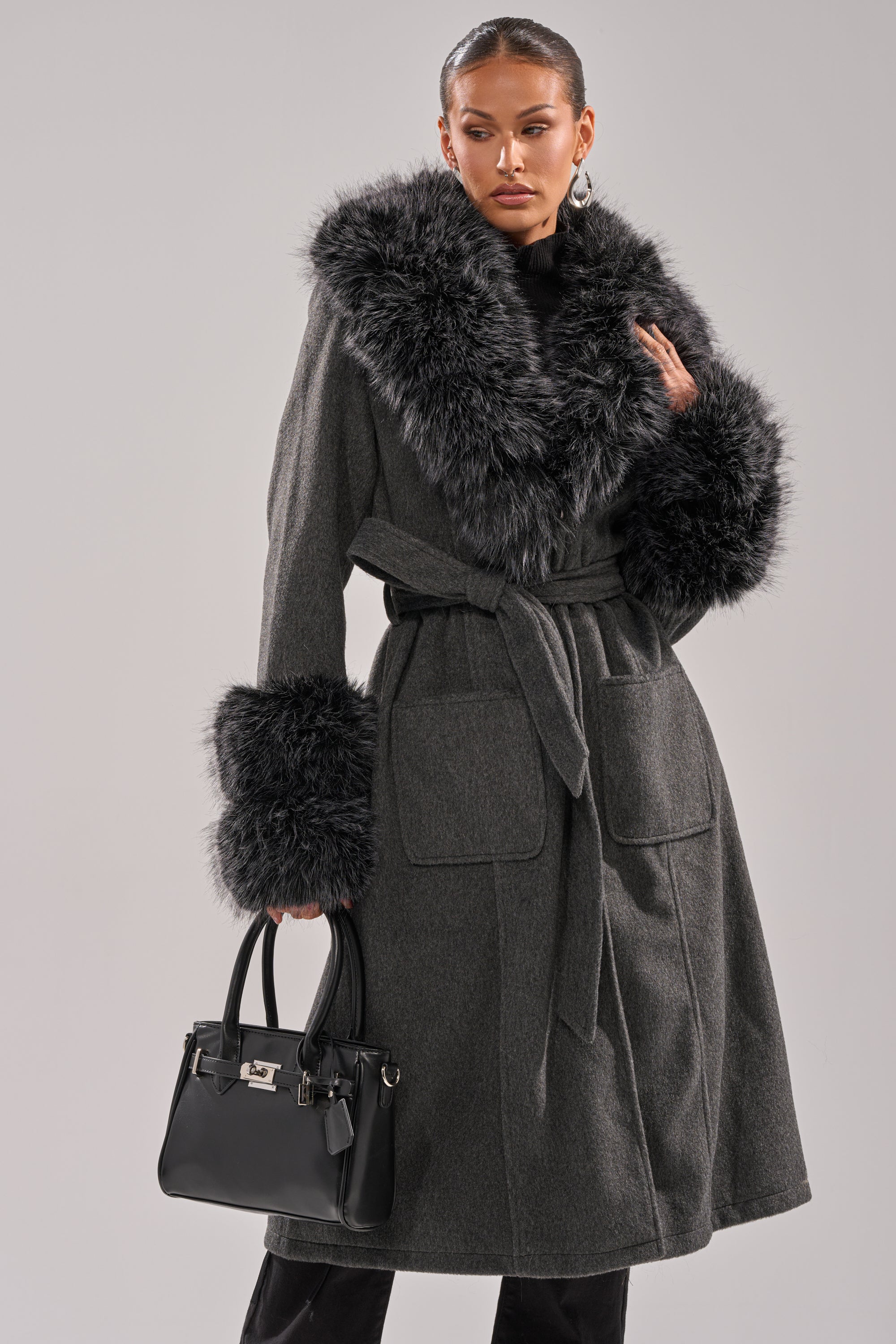 A woman models the HARMONY FUR TRIM TRENCH, a long dark gray winter coat with large faux fur trim on the collar and cuffs, holding a black handbag against a plain light gray background. 0