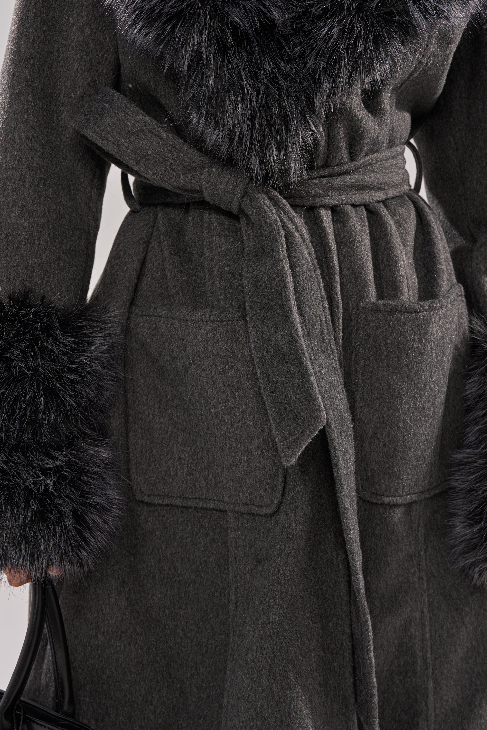 A close-up of the HARMONY FUR TRIM TRENCH in dark gray wool, featuring a waist belt, large front pockets, and black faux fur on the collar and cuffs. The person holds a black handle, highlighting this chic winter jacket. 9