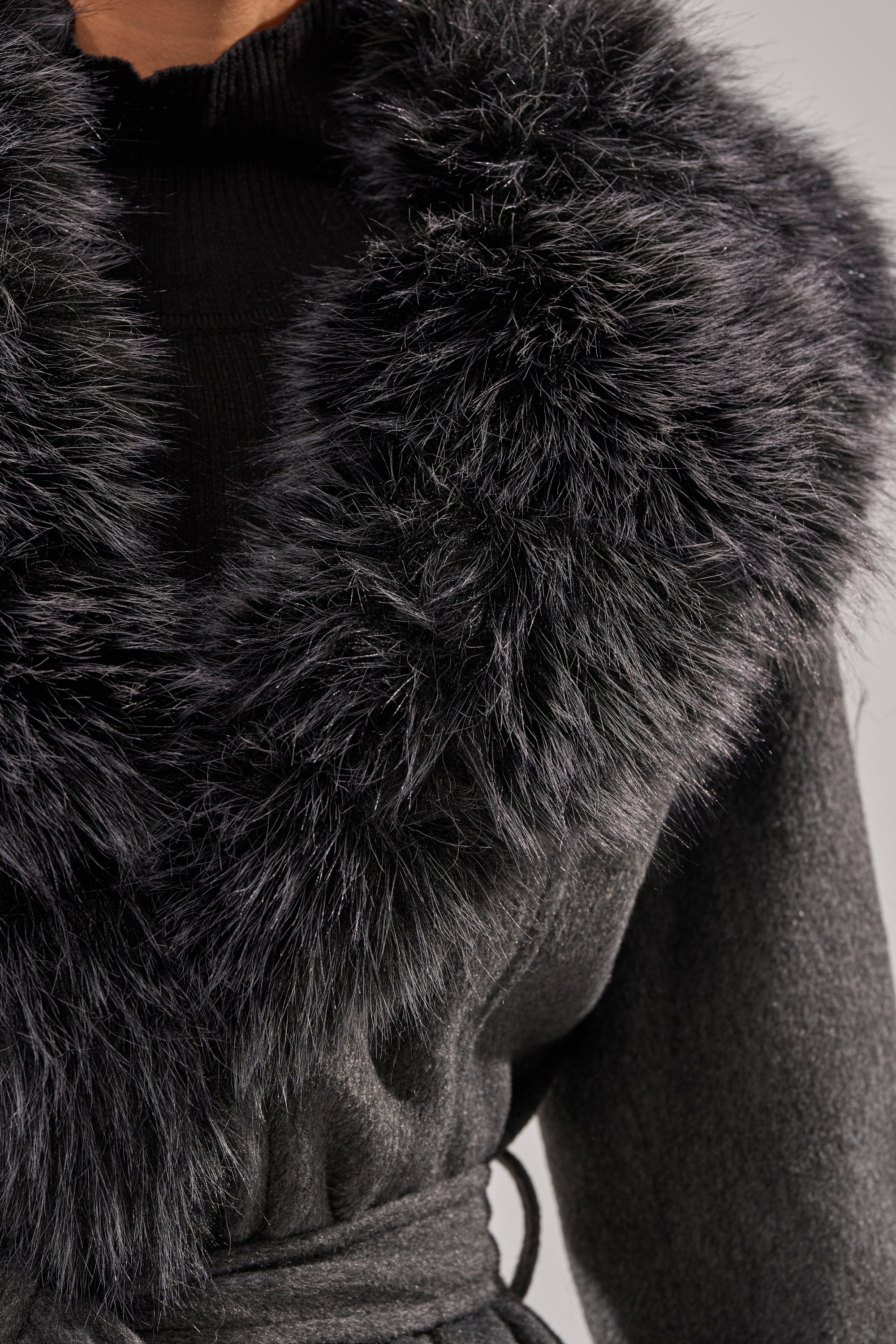 A close-up of a person wearing the HARMONY FUR TRIM TRENCH, a dark wool winter jacket with a large black faux fur collar, layered over a black ribbed turtleneck sweater. 10