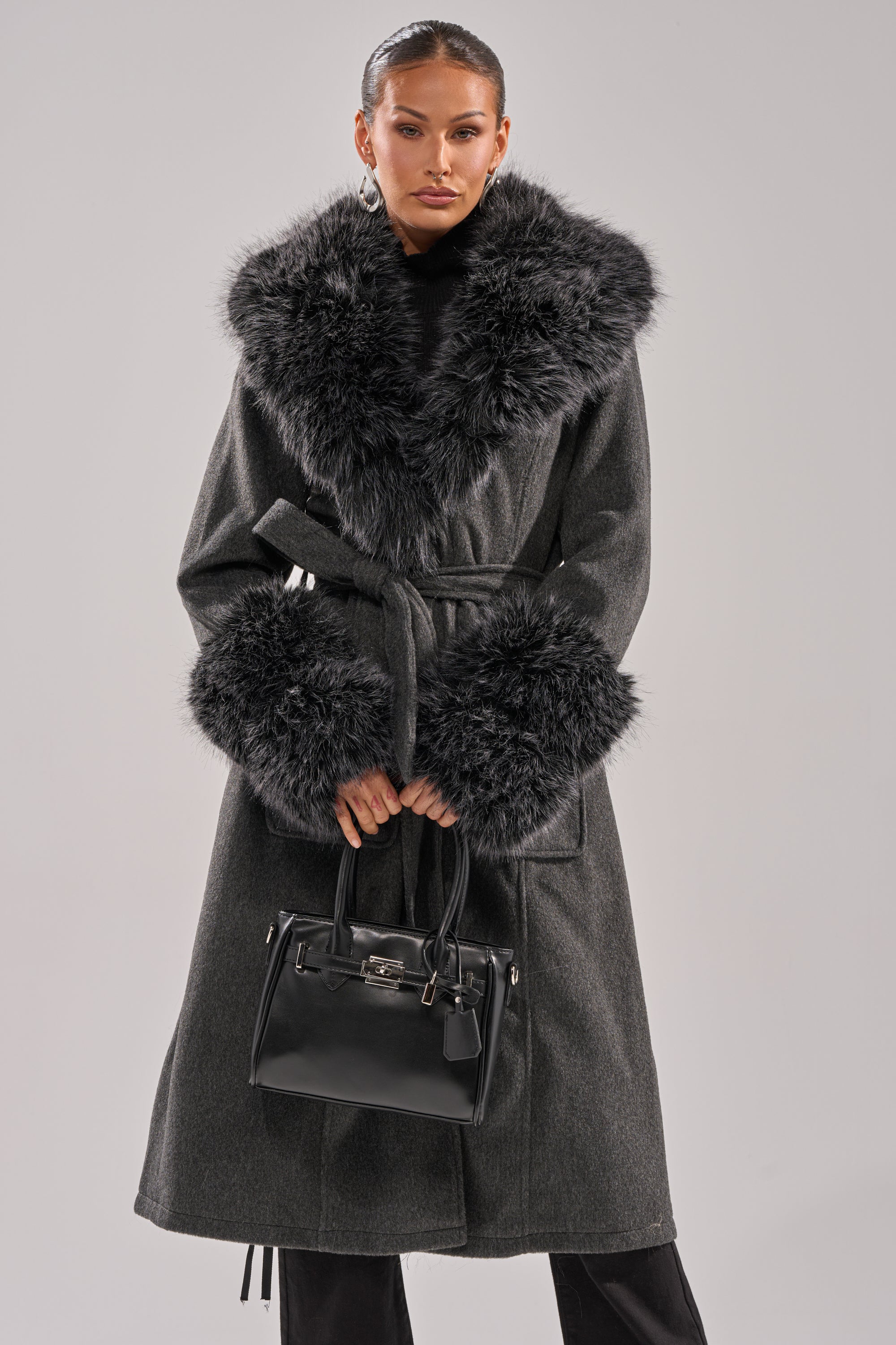 A person wears the HARMONY FUR TRIM TRENCH, a long dark gray winter coat with large black faux fur trim on the collar and cuffs, holding a black handbag against a plain, light background. 2