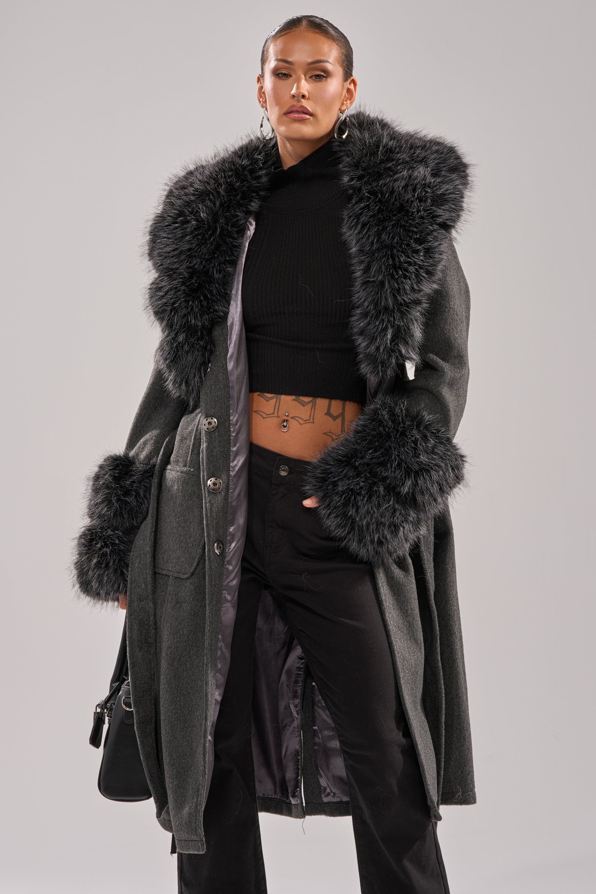 A woman stands confidently, showing her tattooed midriff in the HARMONY FUR TRIM TRENCH, a long winter coat with a large faux fur collar and cuffs, paired with a black cropped turtleneck, black pants, and hoop earrings. 3