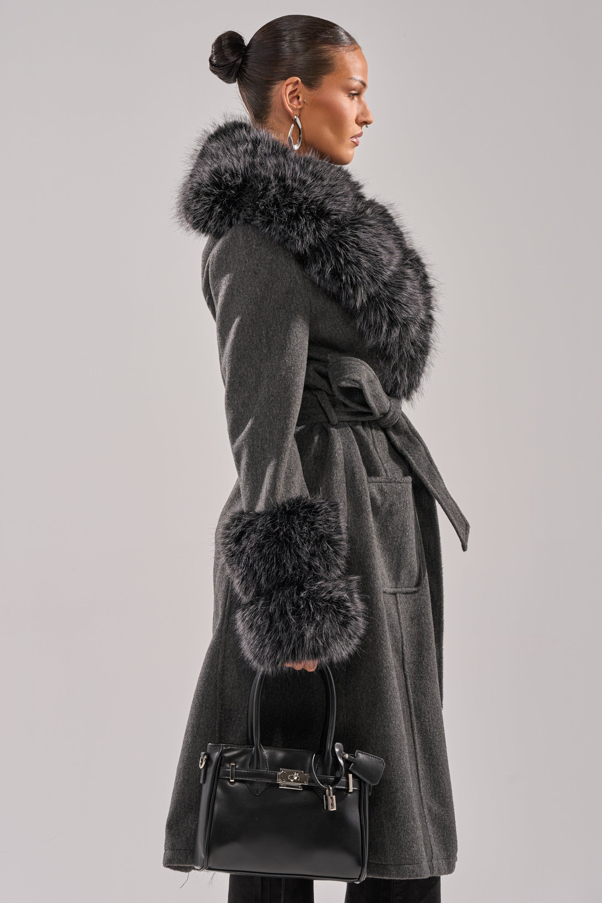 A woman in profile models the HARMONY FUR TRIM TRENCH—a long, belted gray winter coat with large faux fur collar and cuffs—holding a small black handbag, set against a plain light gray background. 5