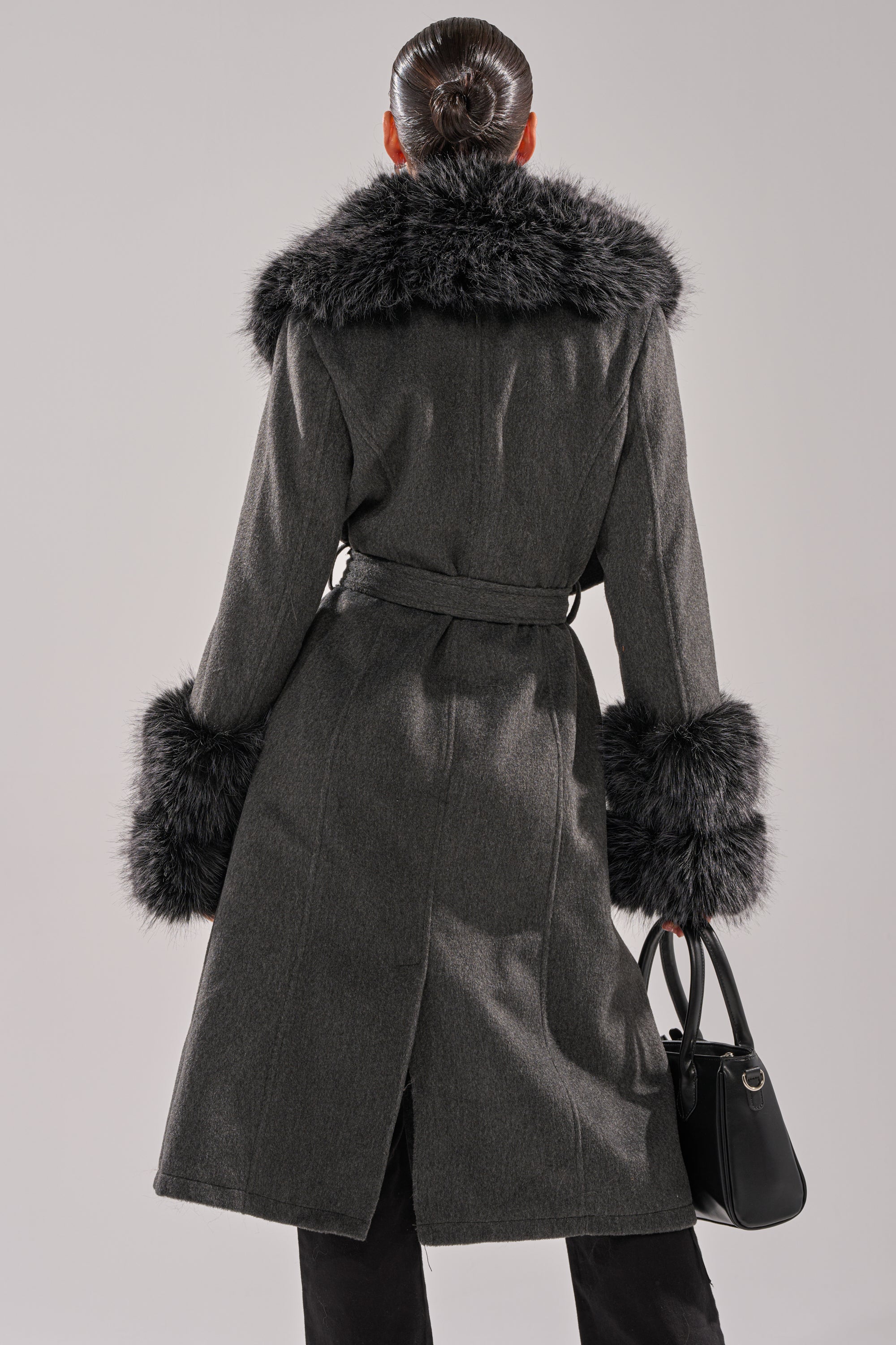 A person with dark hair in a sleek bun wears the HARMONY FUR TRIM TRENCH with bold black faux fur collar and cuffs, holding a black handbag, shown from the back against a plain background. 6