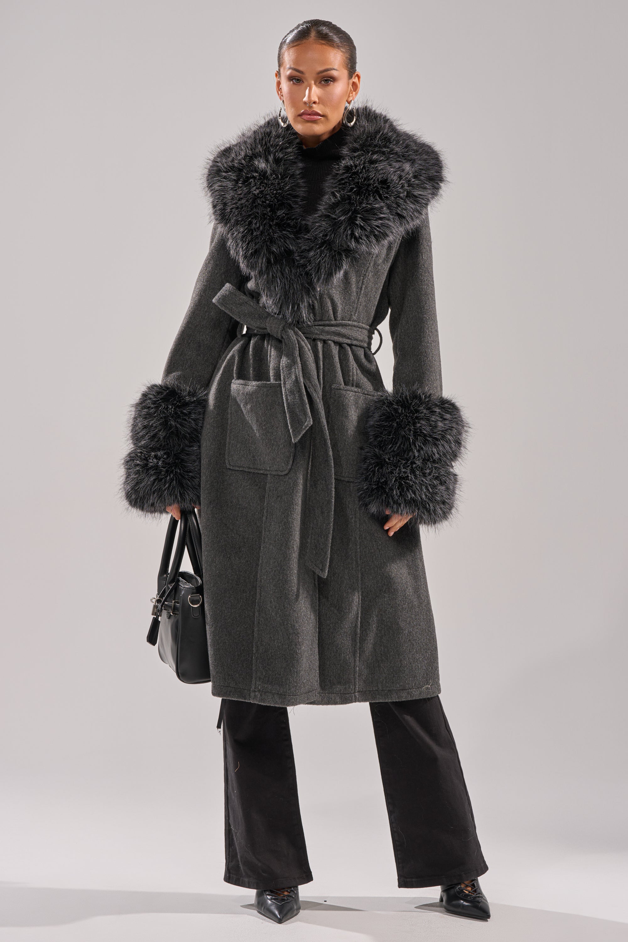 A woman stands against a plain light background wearing the HARMONY FUR TRIM TRENCH, featuring a long dark gray coat with a belted waist, large faux fur collar and cuffs, black pants, pointed shoes, and a black handbag. 7