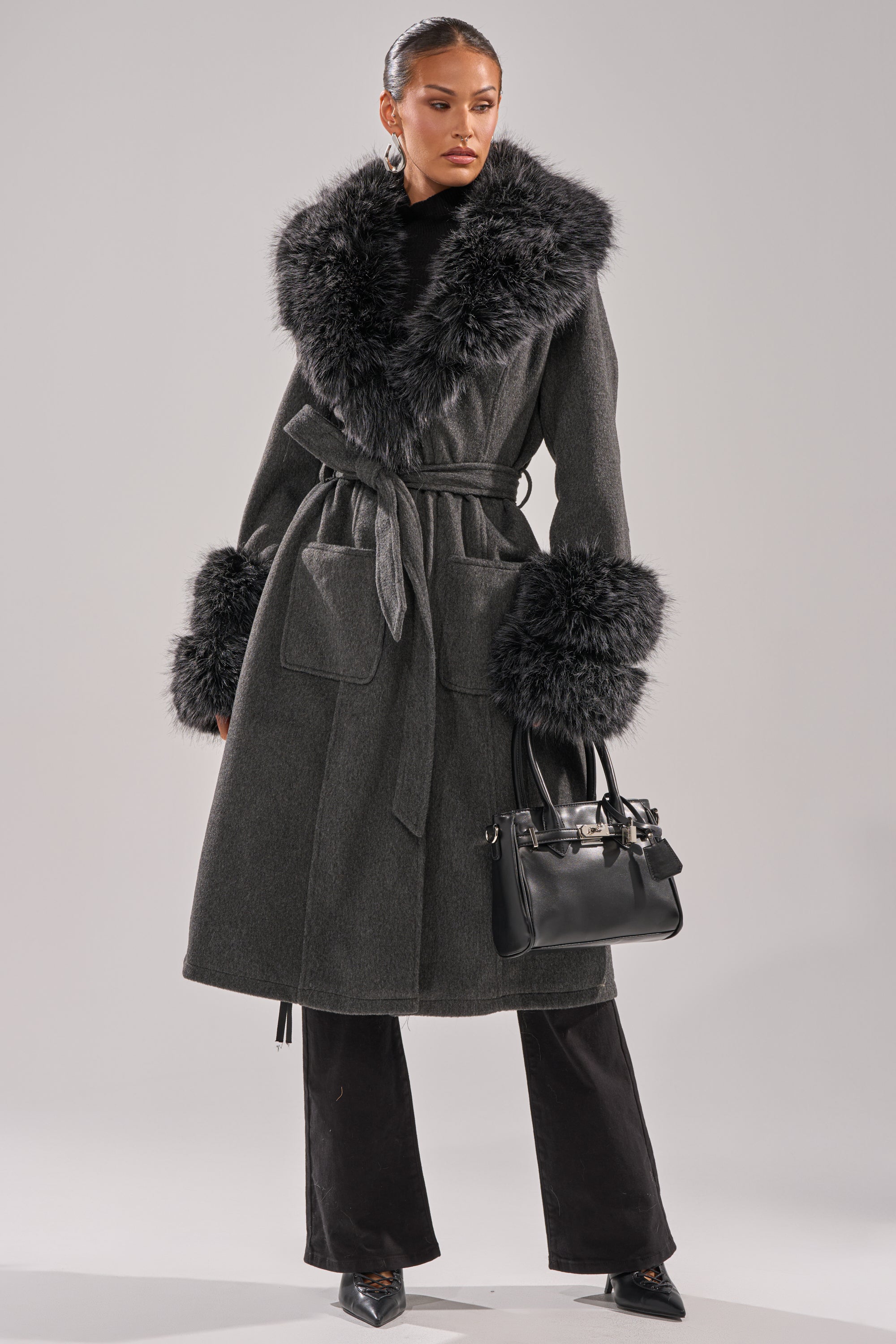 A woman poses in the HARMONY FUR TRIM TRENCH, a long dark gray winter coat featuring large faux fur trim on the collar and cuffs. She holds a black handbag, wears black pants and pointed shoes, set against a plain light background. 8