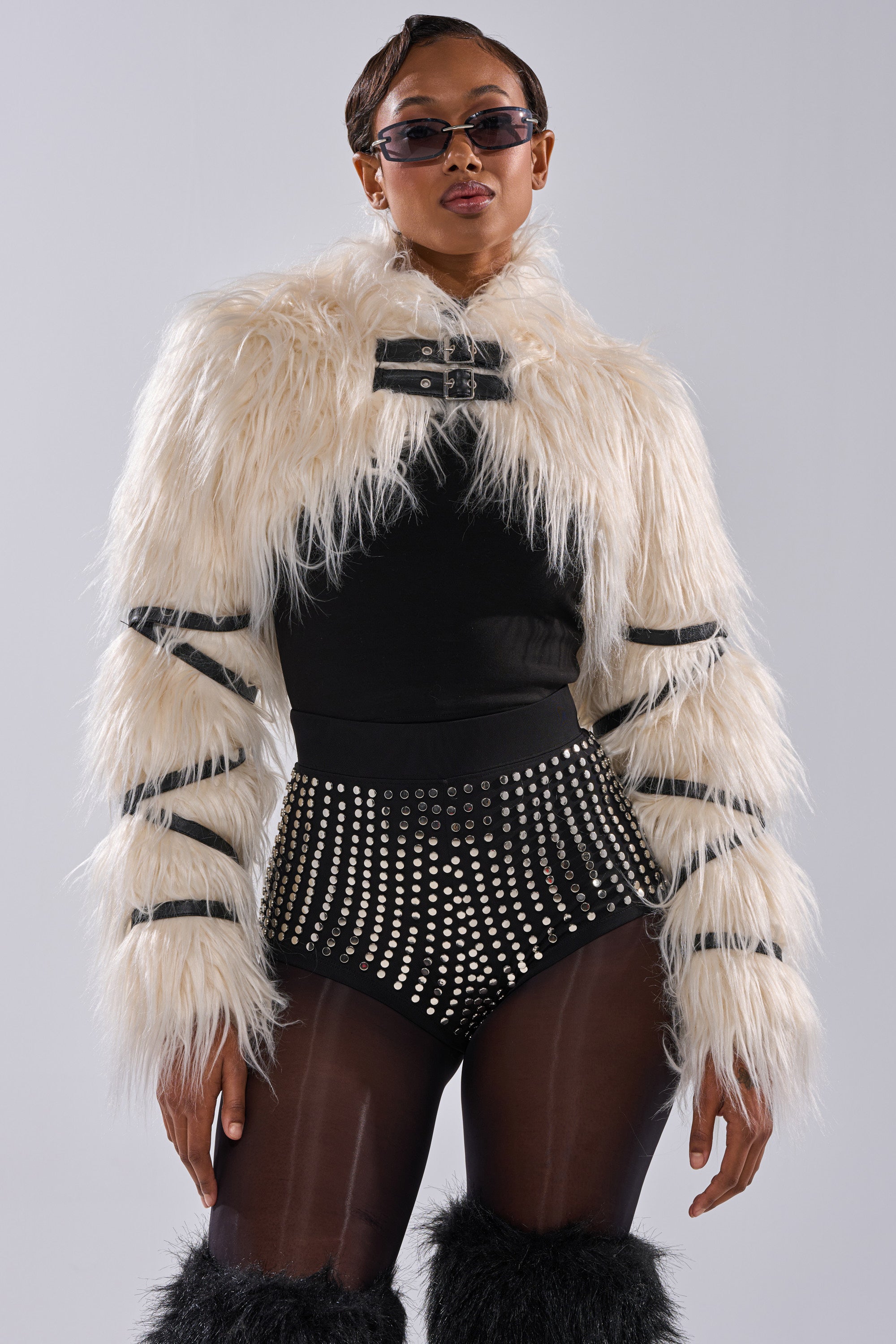 A person poses confidently in the PHIA FAUX FUR BOLERO as a statement piece, styled with black high-waisted shorts featuring metallic studs, sheer black tights, black furry leg warmers, and dark sunglasses.