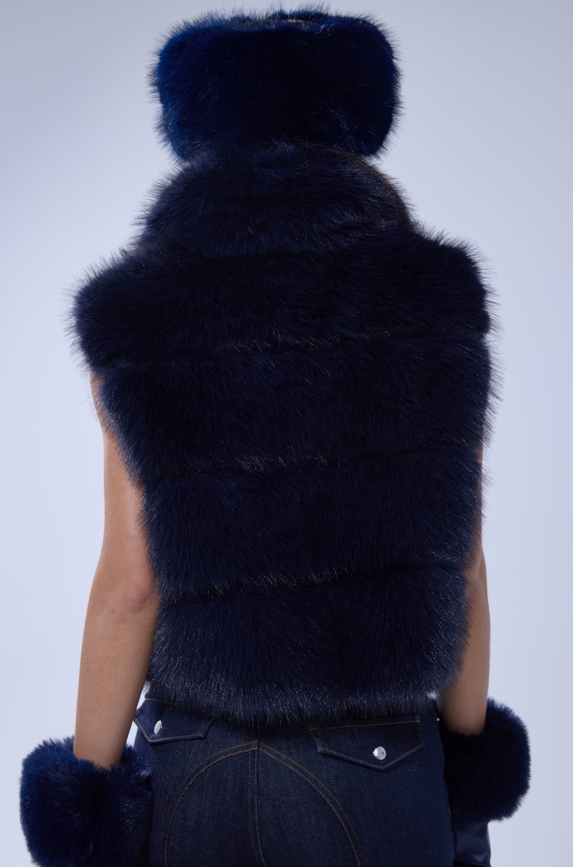 A woman is seen from behind wearing the FAUX FUR VEST IN NAVY, paired with a matching fur hat, fur-trimmed gloves, and dark jeans against a plain light background.