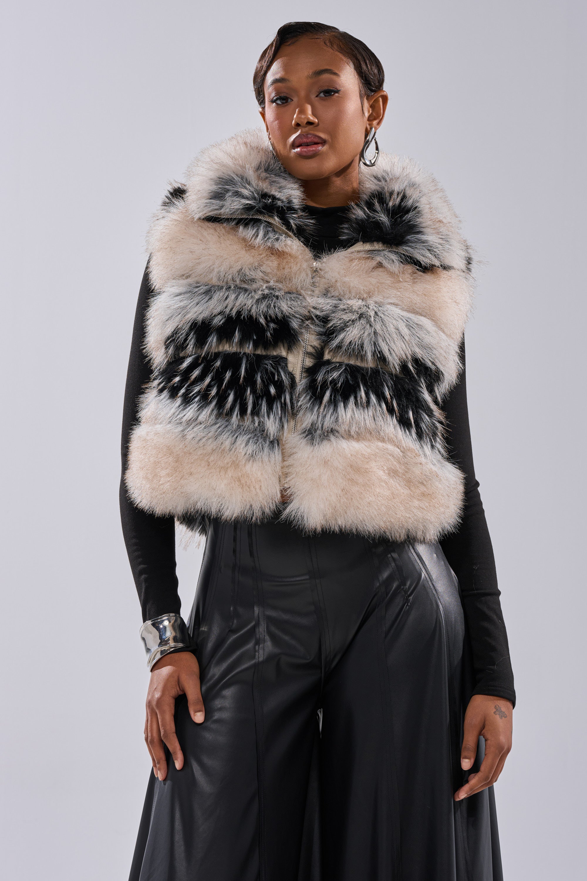 A woman with short hair poses confidently against a gray background in a boxy black long-sleeve top, wide-leg pants, a chunky silver bracelet, and the KIKI FAUX FUR VEST featuring a large, fluffy, multi-colored faux fur and oversized collar.