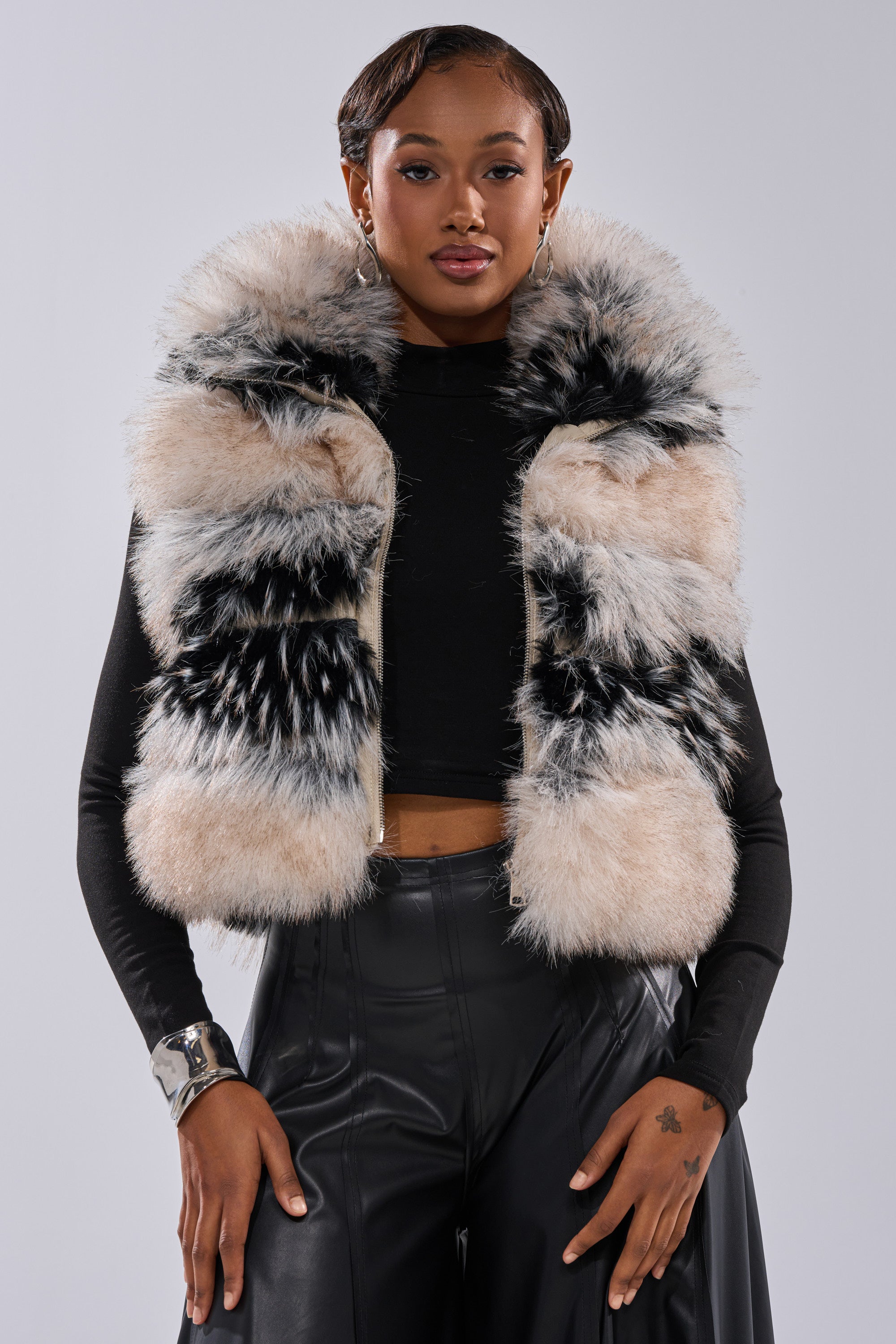 A woman faces forward wearing the KIKI FAUX FUR VEST—a sleeveless striped faux fur vest with beige and black patterns—styled over a black long-sleeve top and black leather pants, accessorized with a silver cuff bracelet and small earrings.