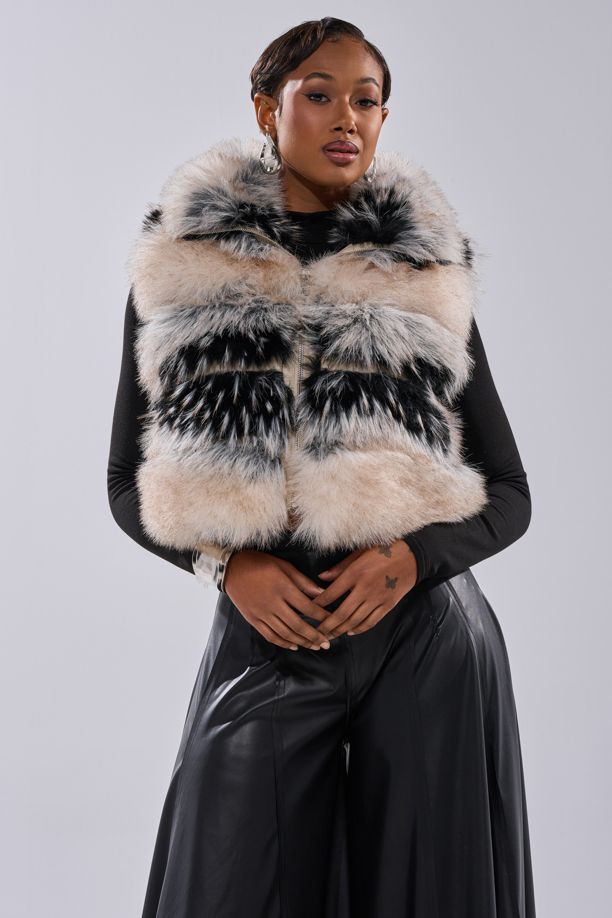 A woman models the KIKI FAUX FUR VEST—fluffy, black and white—over a black long-sleeve top and wide-leg pants, posing confidently against a plain light gray background.