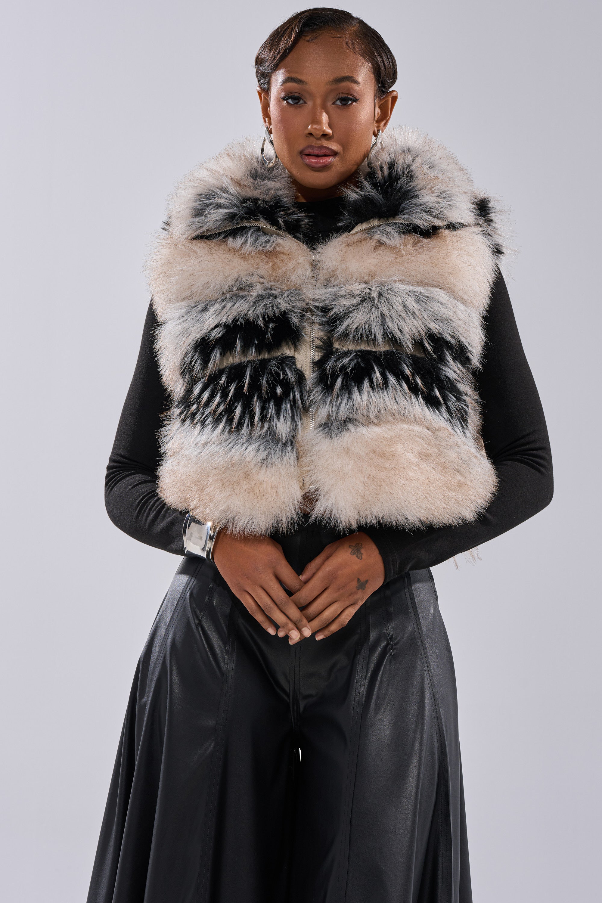 A woman stands against a light gray background wearing the KIKI FAUX FUR VEST, a fluffy multicolored piece with an oversized collar, over a black long-sleeve top and black wide-leg pants, posing with her hands clasped in front of her.