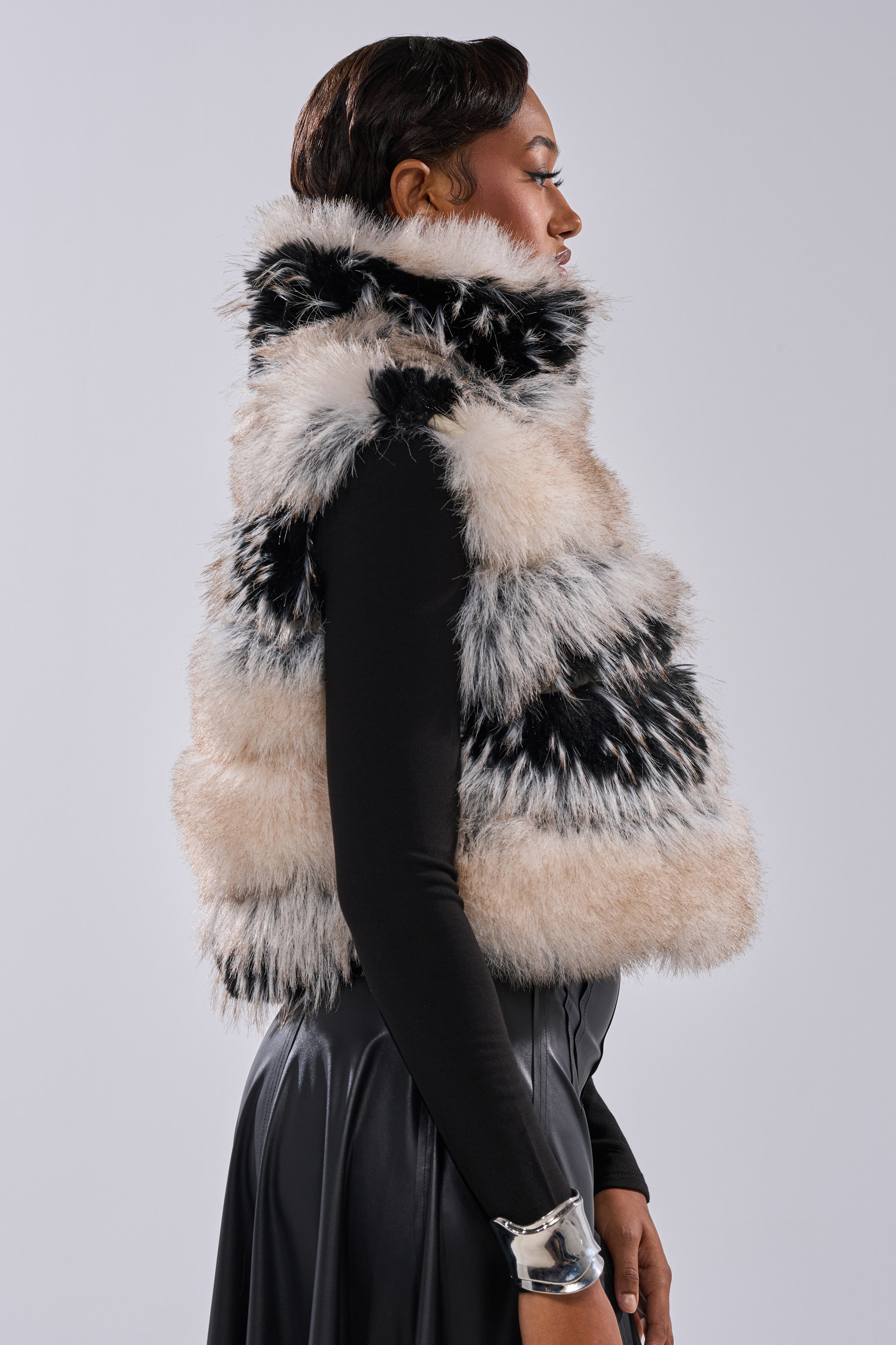 A person in profile wears the KIKI FAUX FUR VEST—a sleeveless, striped black and white piece—over a black long-sleeve top and skirt, accessorized with a chunky silver bracelet, set against a plain light gray background.