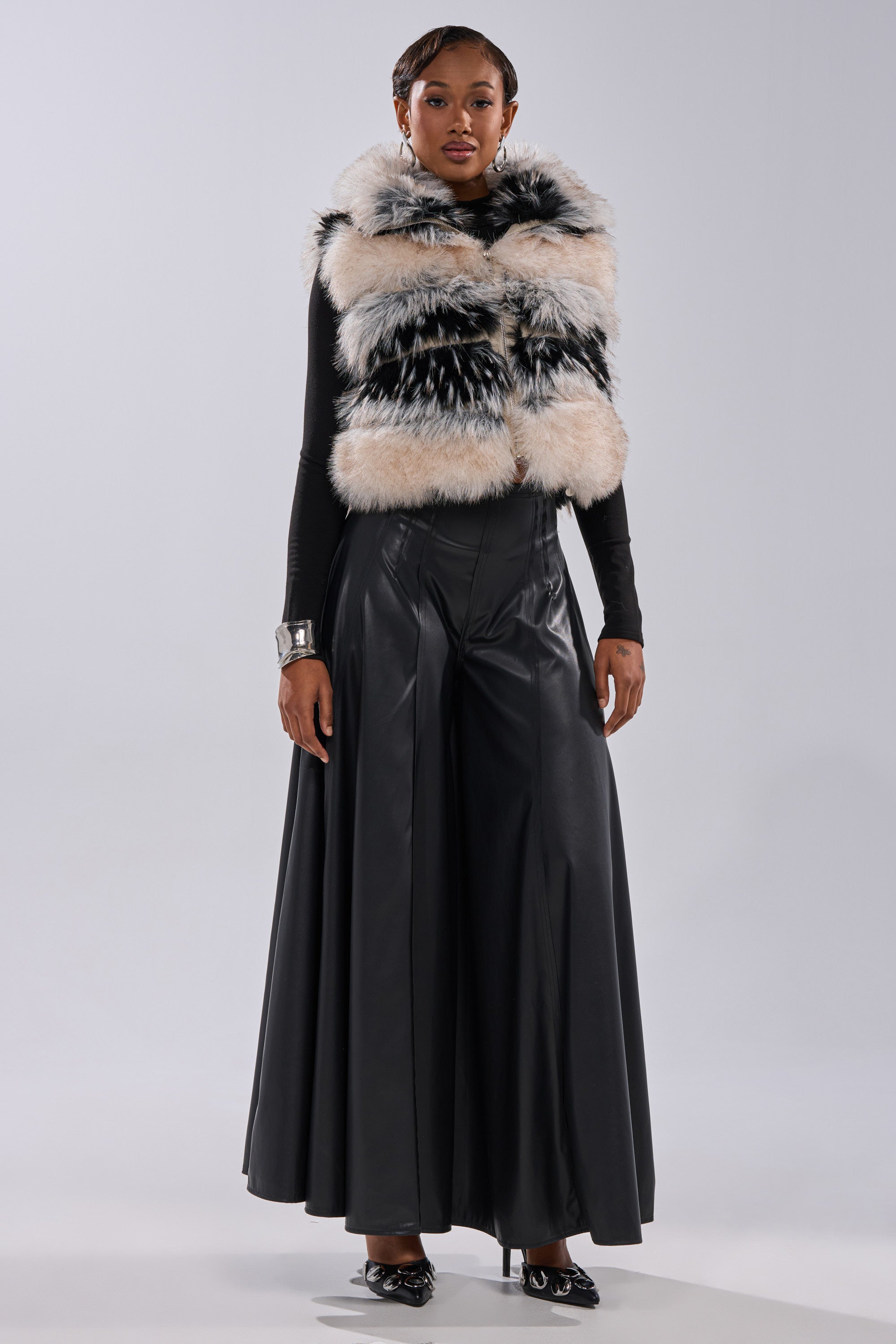A woman stands facing forward wearing a black long-sleeve top, wide-leg black leather pants, pointed black shoes, a large silver cuff bracelet, and the KIKI FAUX FUR VEST.