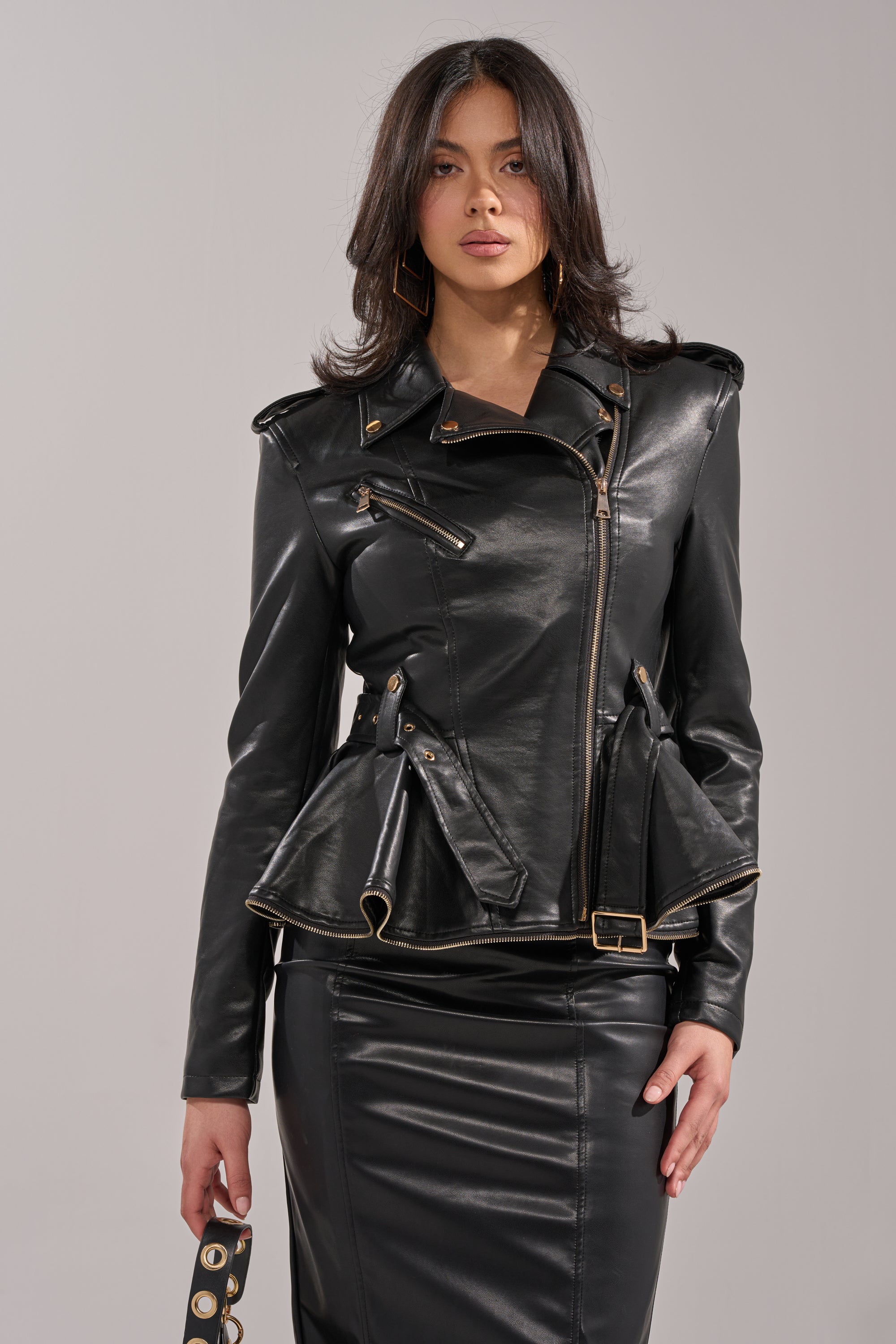 A woman with shoulder-length dark hair faces forward in the JULES PEPLUM MOTO, a chic black faux leather jacket with gold zippers, paired with a matching skirt. She holds a strap in her left hand against a plain, light background. 0