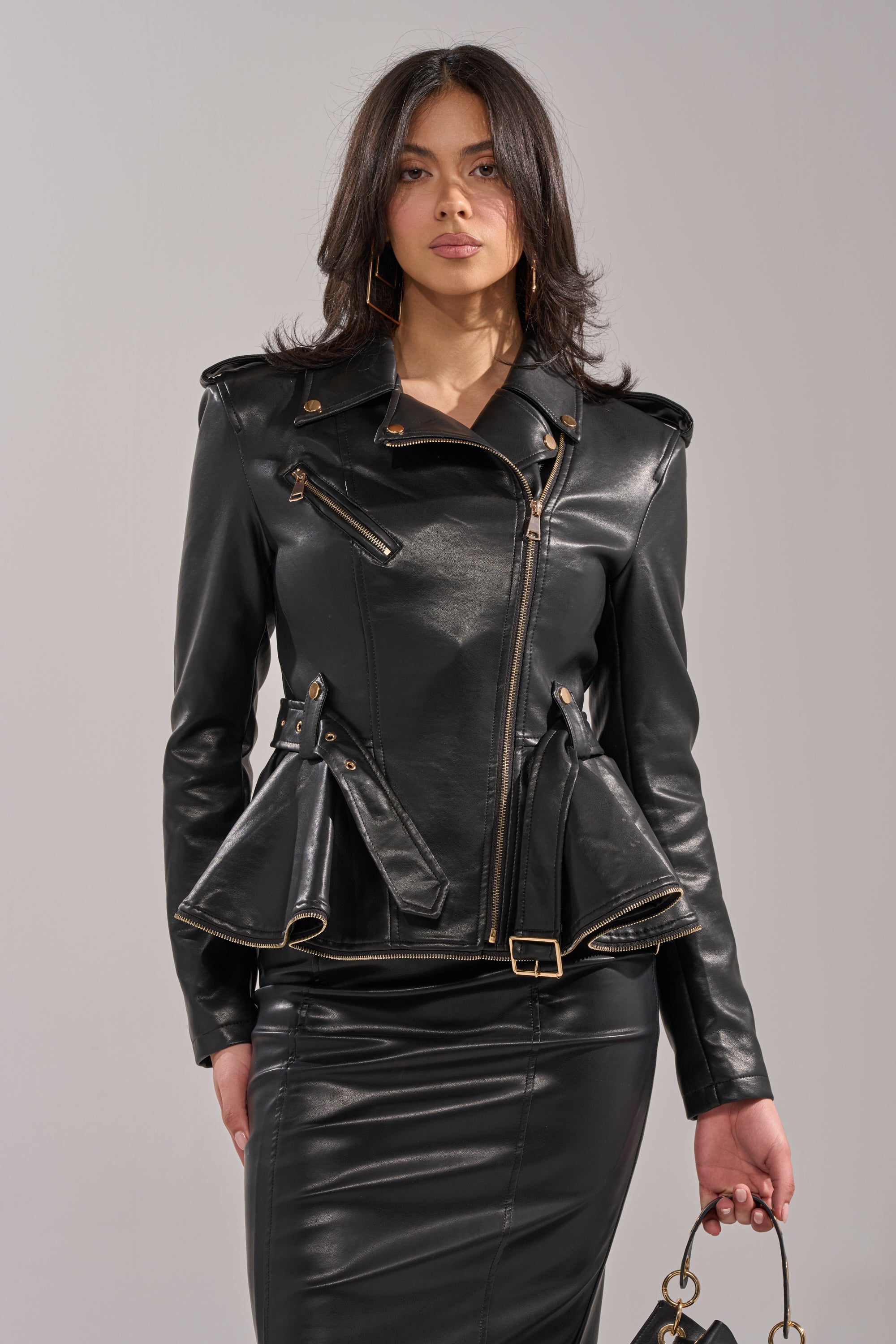 A woman poses against a plain light background holding a handbag, dressed in the JULES PEPLUM MOTO fitted black faux leather jacket with gold zippers and accents, paired with a matching black leather skirt. 1