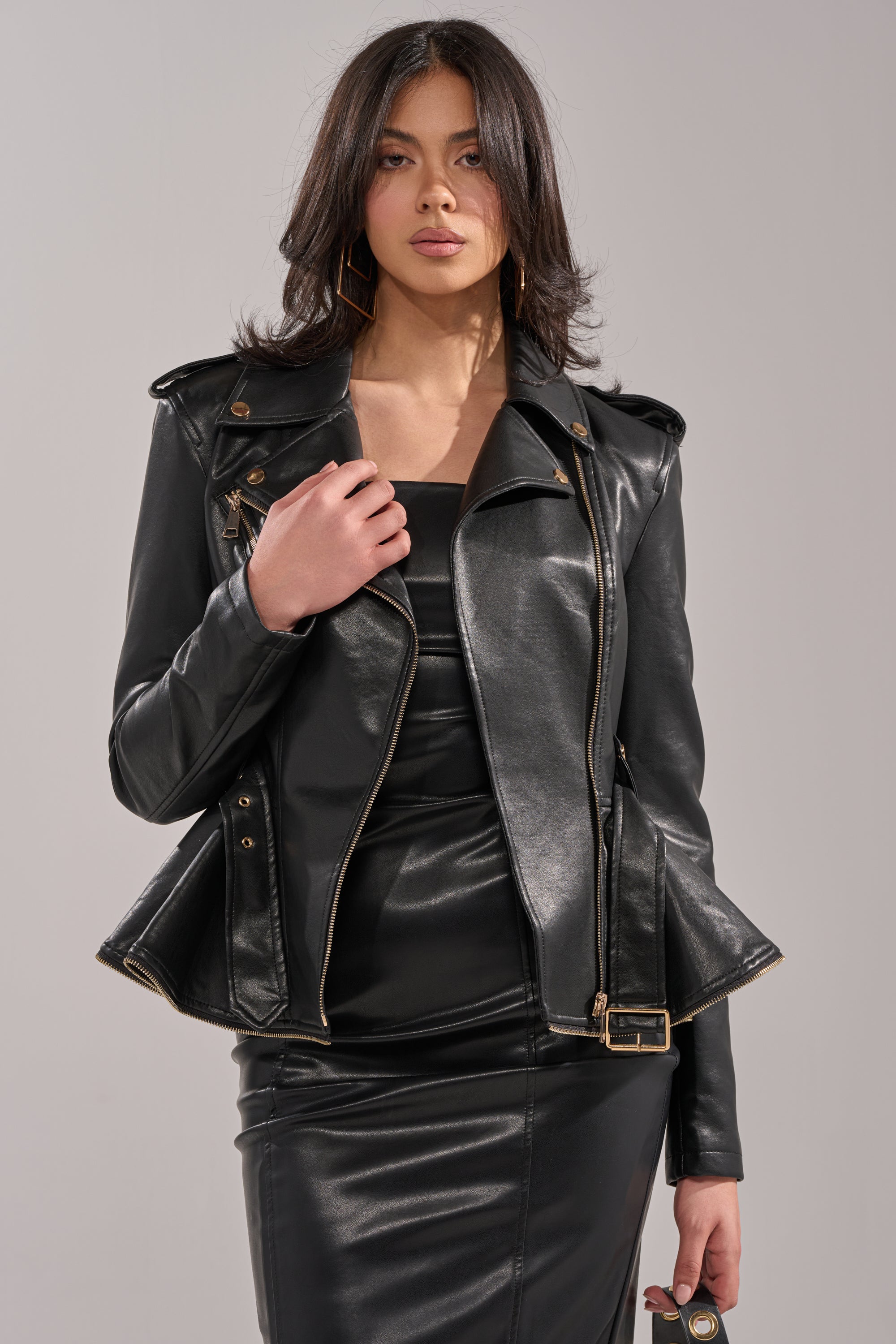 A woman with wavy dark hair wears the JULES PEPLUM MOTO jacket over a black dress, standing confidently against a plain light gray background and looking at the camera. 2