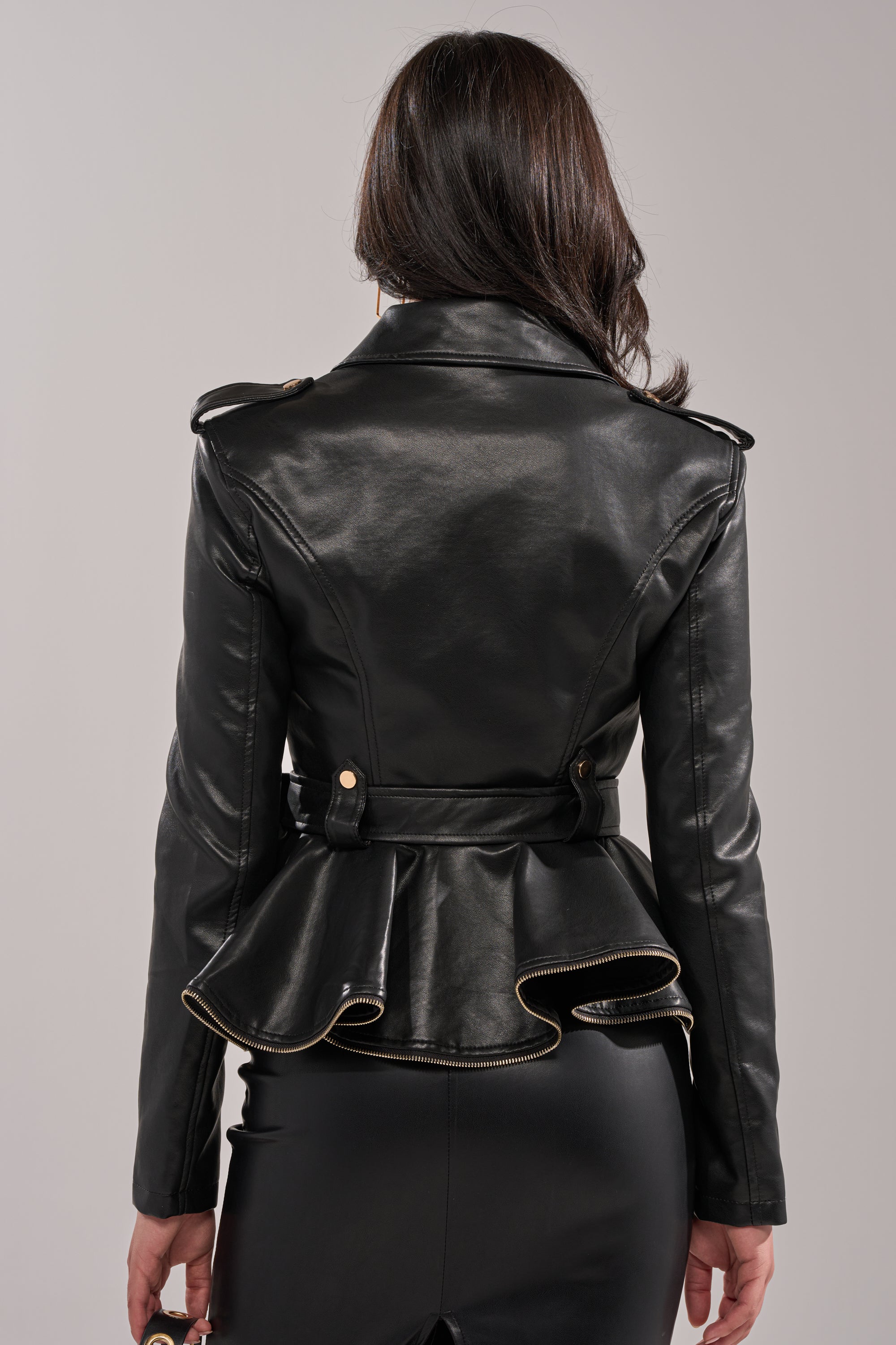 A woman with dark hair, seen from behind, is wearing the JULES PEPLUM MOTO—a black faux leather moto jacket with a flared peplum hem and gold button details—paired with a black skirt. 6